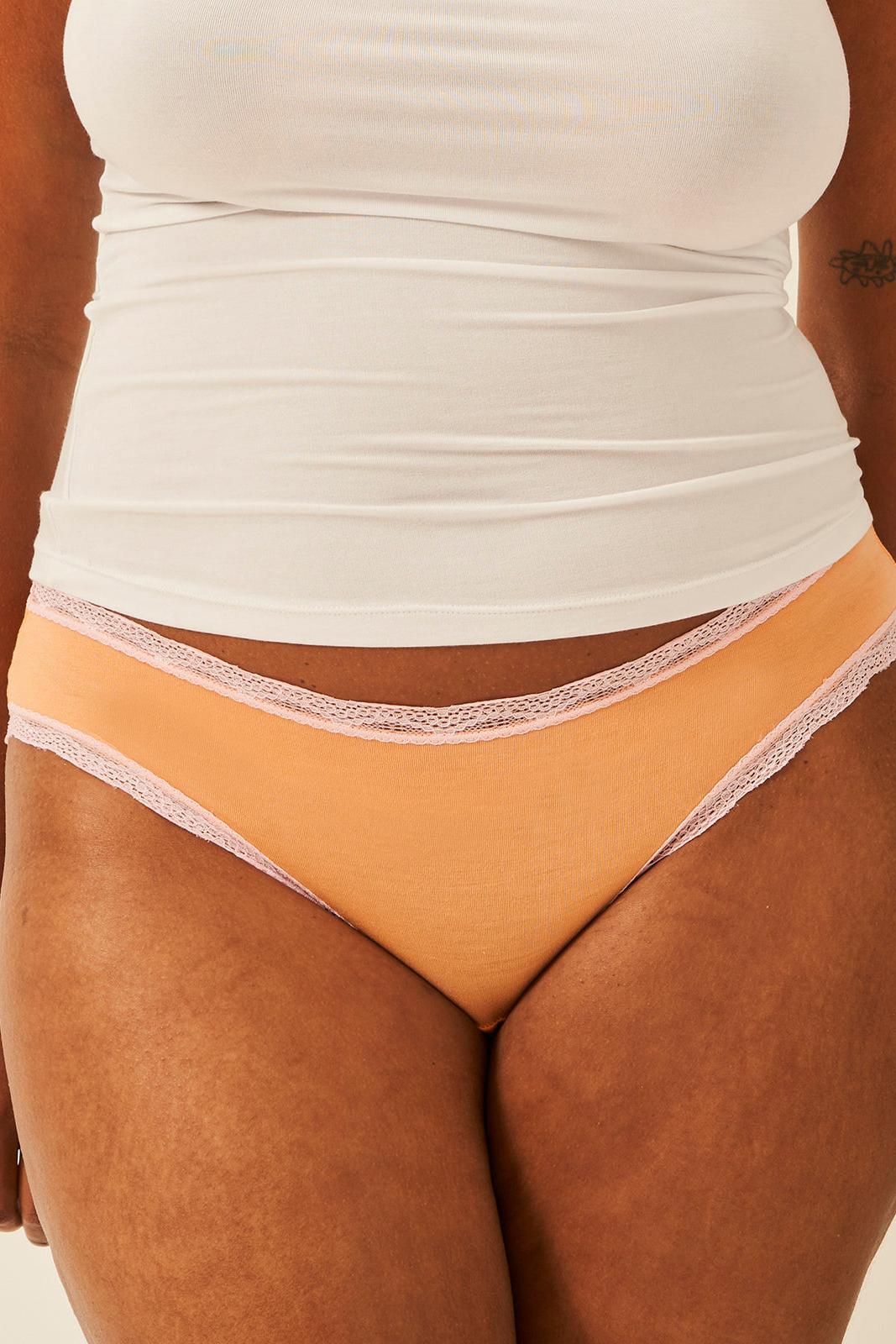 A close-up of a model wearing a peach-colored high-cut brief with light pink lace trim, paired with a white tank top.