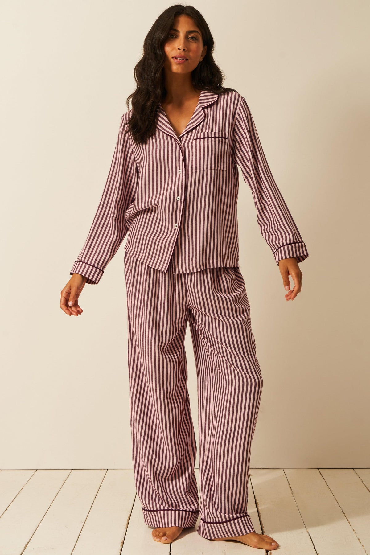 A woman with dark hair standing in a button-down pyjama set with vertical stripes against a plain beige background.