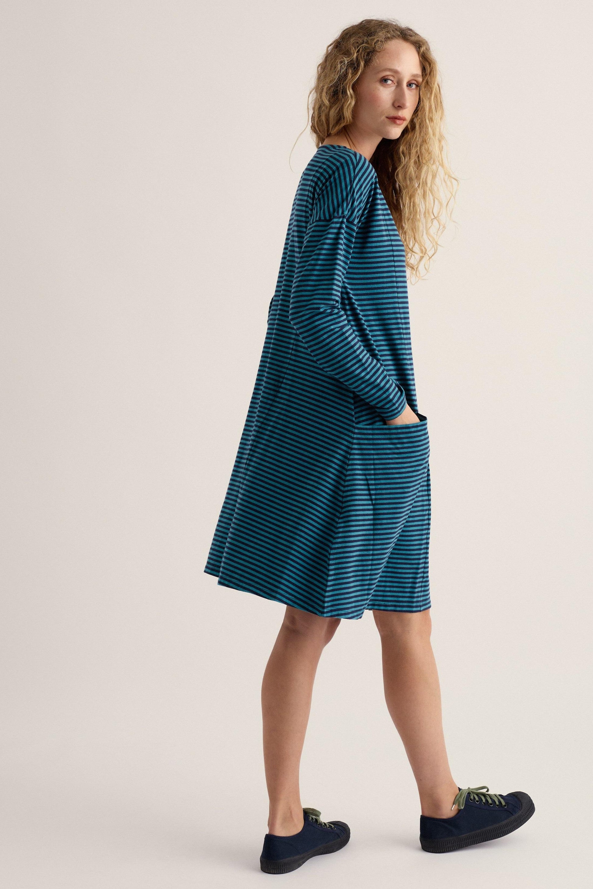 A woman modeling a teal blue and white striped long-sleeve dress with side pockets, paired with dark navy loafers, against a solid light beige studio background.