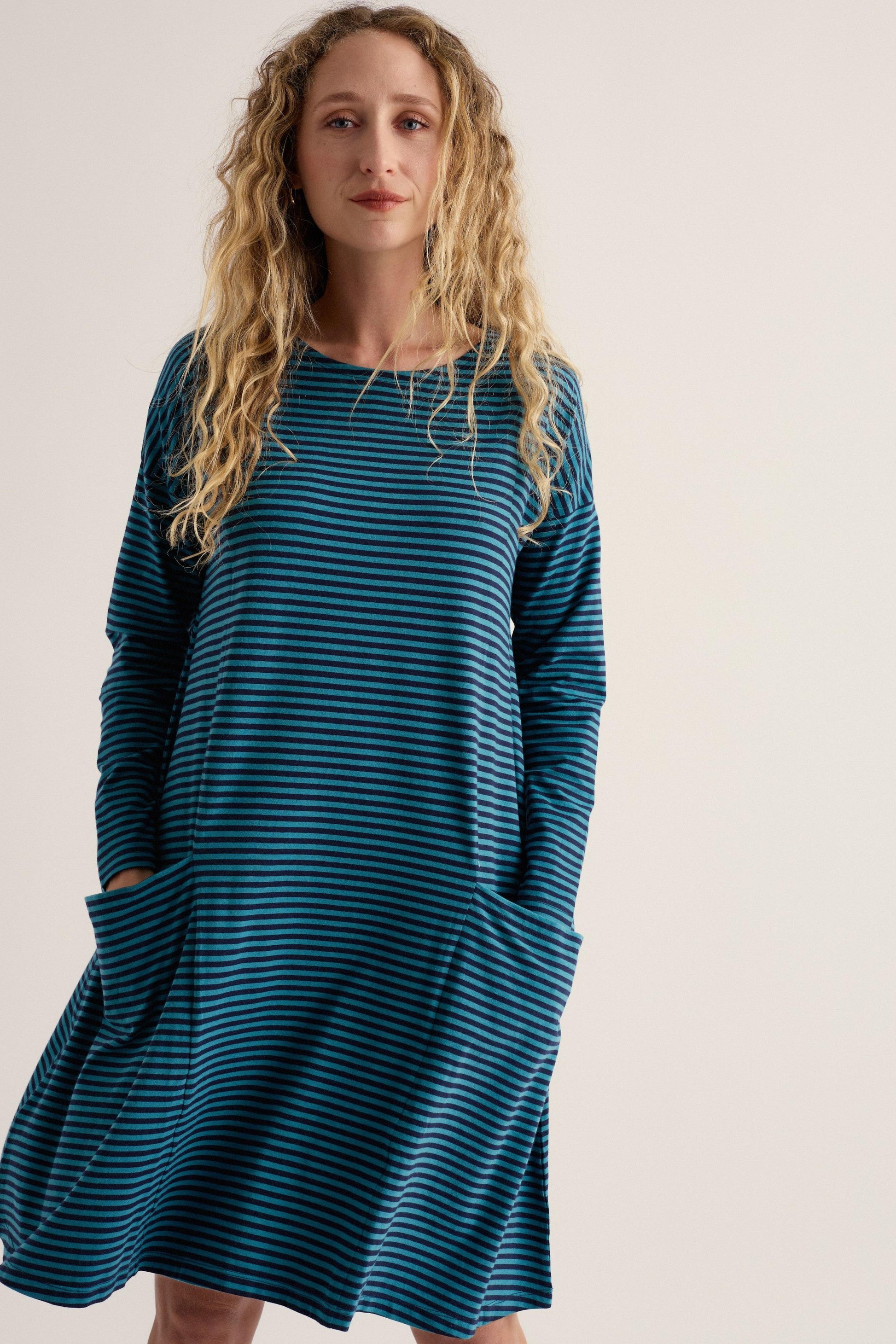 A woman with long curly blonde hair wearing a dark teal blue and black striped long-sleeve dress with side pockets, standing against a plain light beige background.