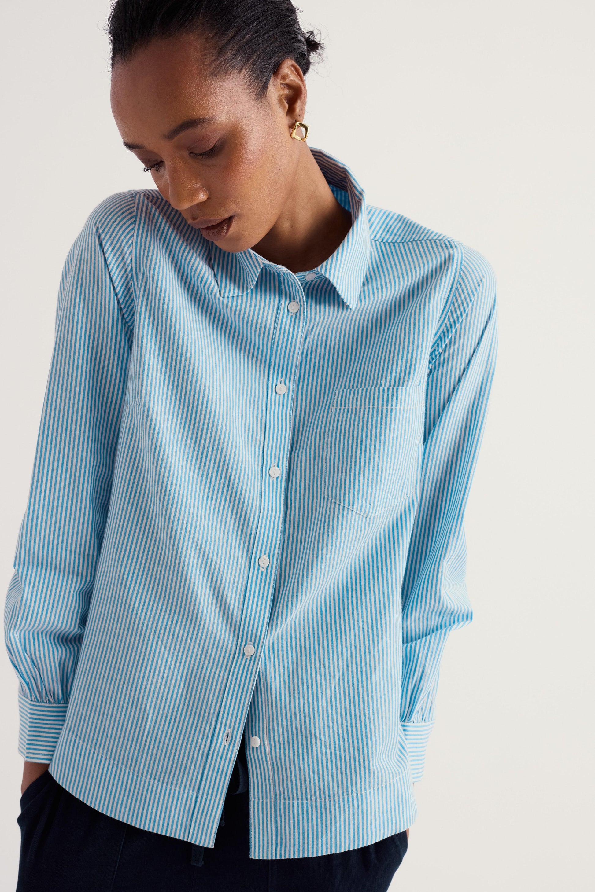 A woman wearing a light blue and white vertically striped button-down shirt, shown from the chest down against a plain light beige studio background.