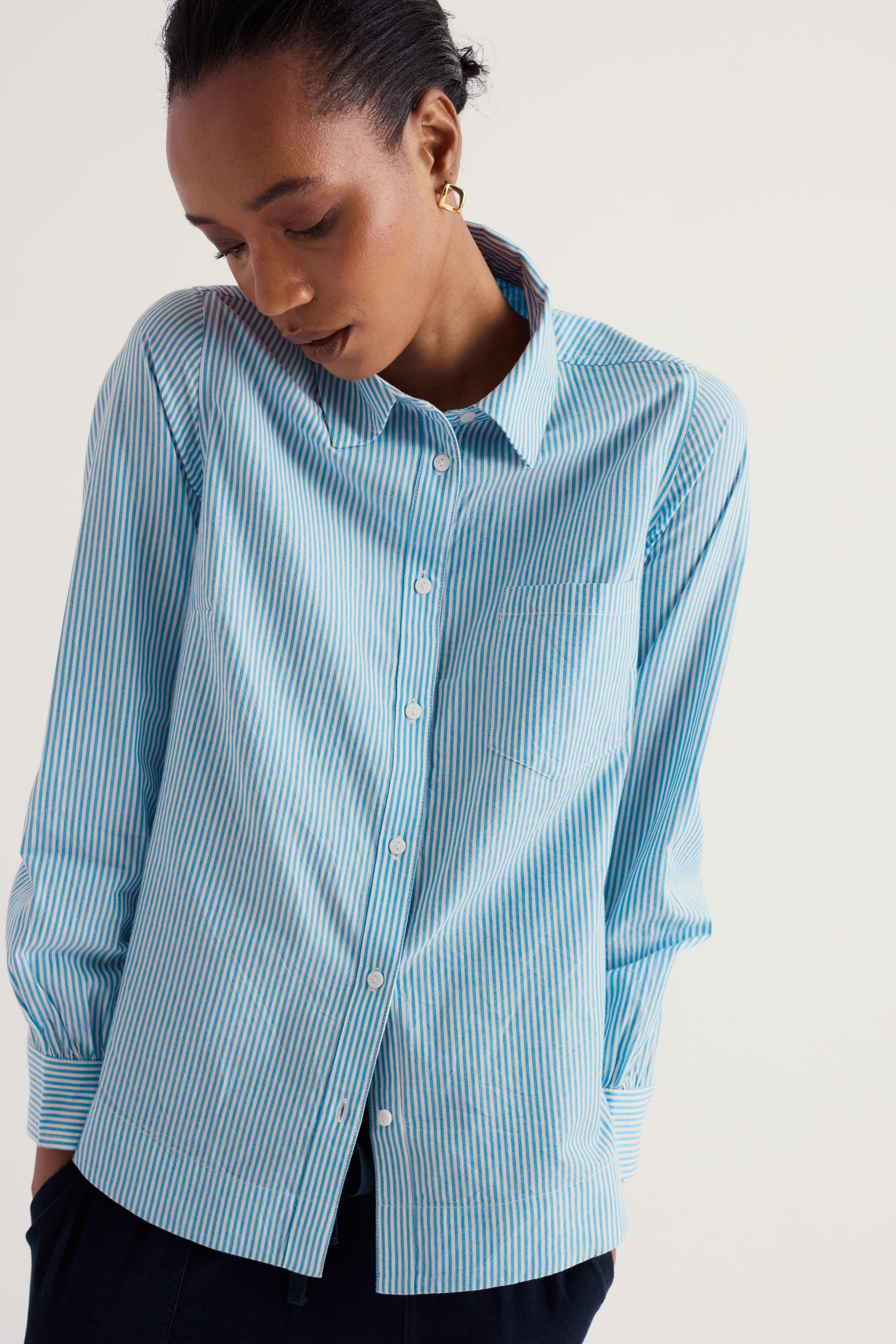 A woman wearing a light blue and white vertically striped button-down shirt, shown from the chest down against a plain light beige studio background.