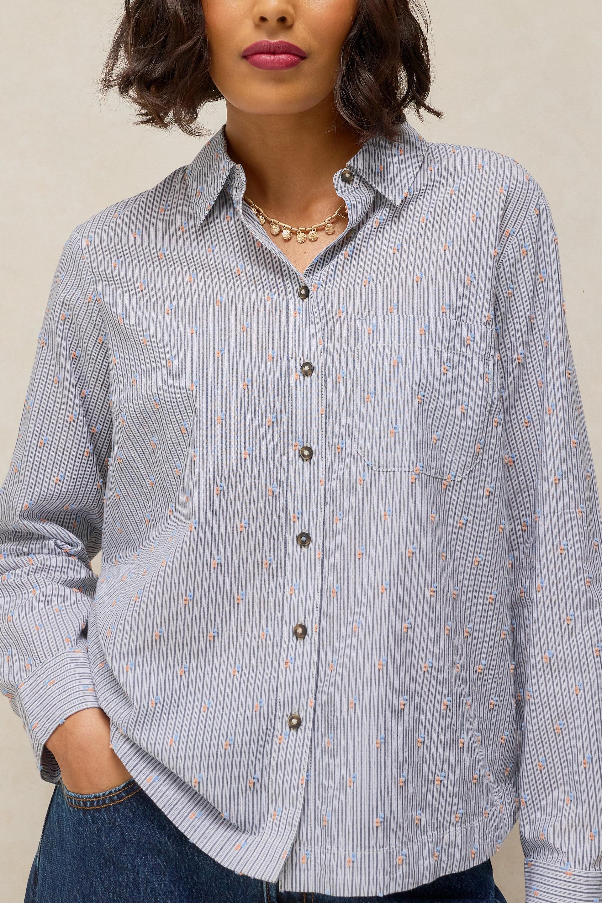 A woman wearing a light blue and white vertically striped button-up shirt with small embroidered details, paired with dark blue jeans and a gold necklace, against a neutral beige background.