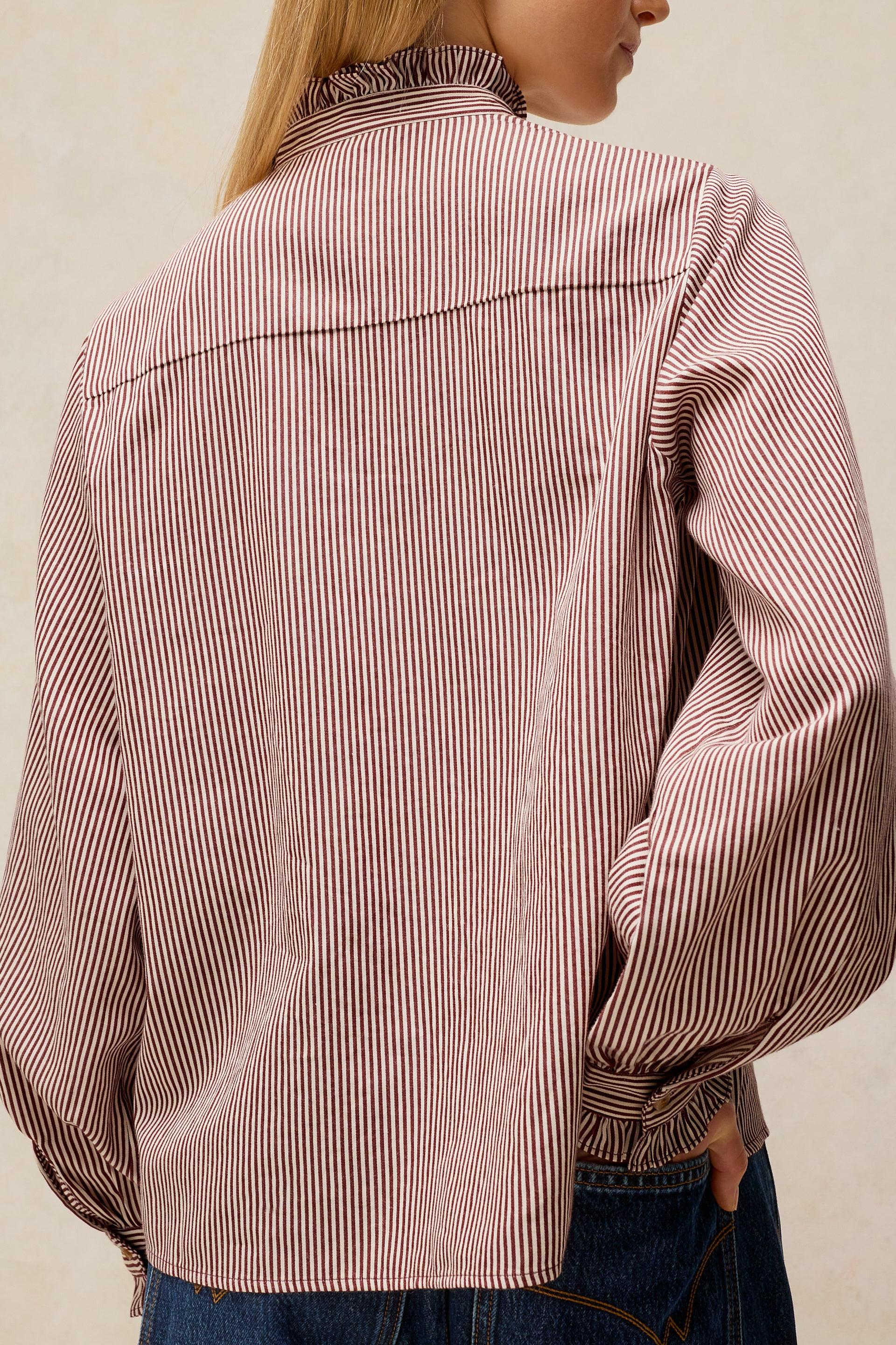 A person shown from the back wearing a long-sleeve button-up shirt with thin vertical pink and white stripes, paired with blue jeans against a plain light beige background.