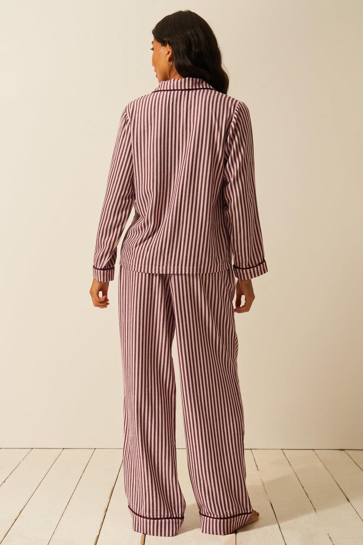 A woman stands facing away, wearing a long-sleeved button-up shirt and matching pyjama pants with a dark purple stripe pattern.