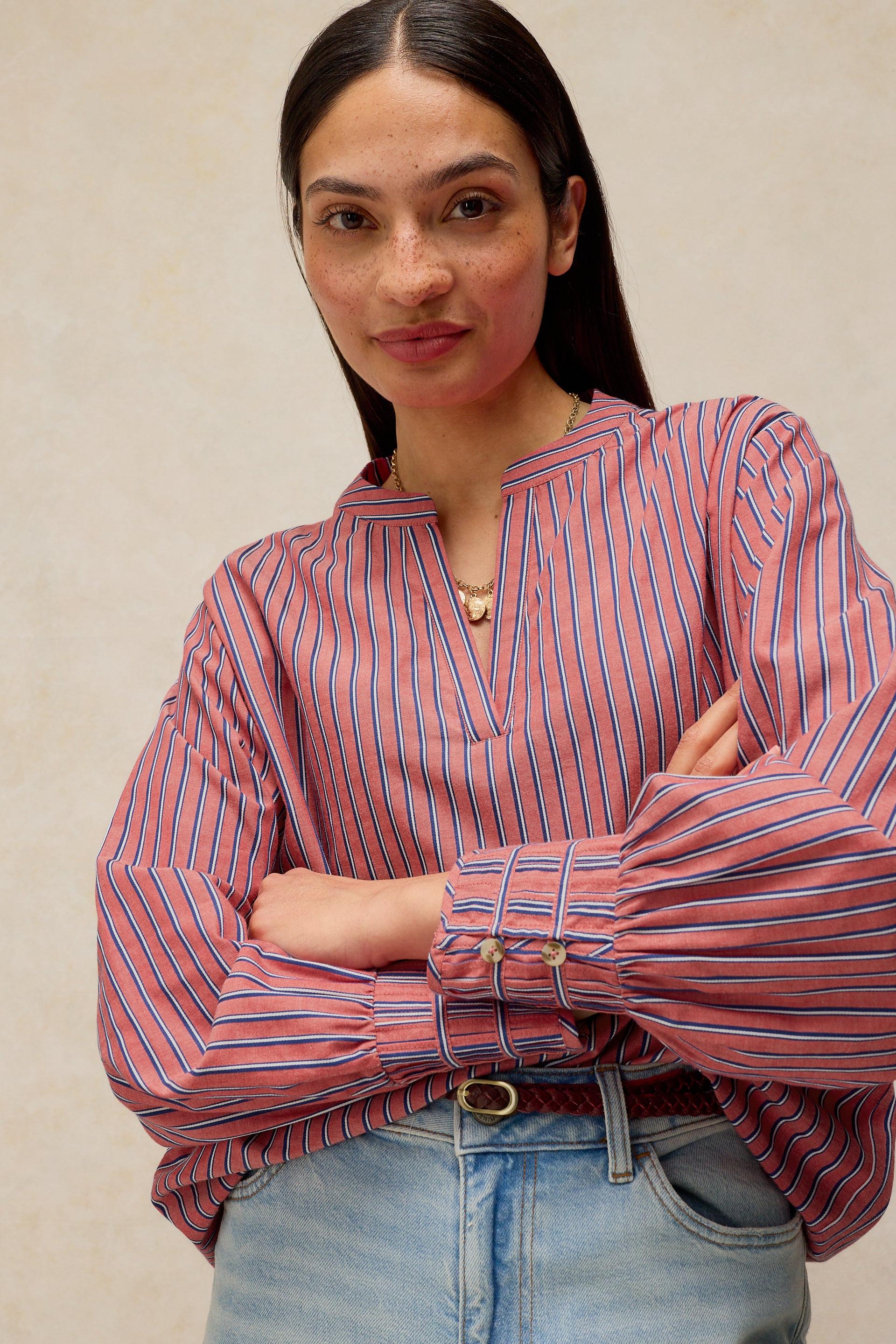 A woman wearing a pink and blue vertically striped long-sleeve blouse with a split neckline and gathered cuffs, paired with light blue jeans, standing against a neutral beige background.