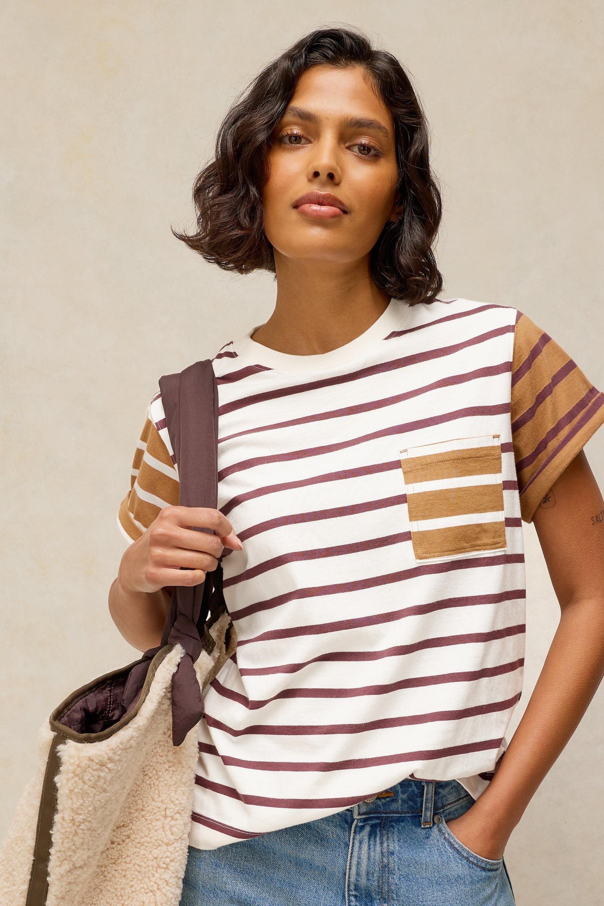 A woman wearing a short sleeve white t-shirt with brown horizontal stripes, a brown chest pocket, and brown striped short sleeves, paired with blue jeans and holding a beige and brown shoulder bag against a neutral beige background.