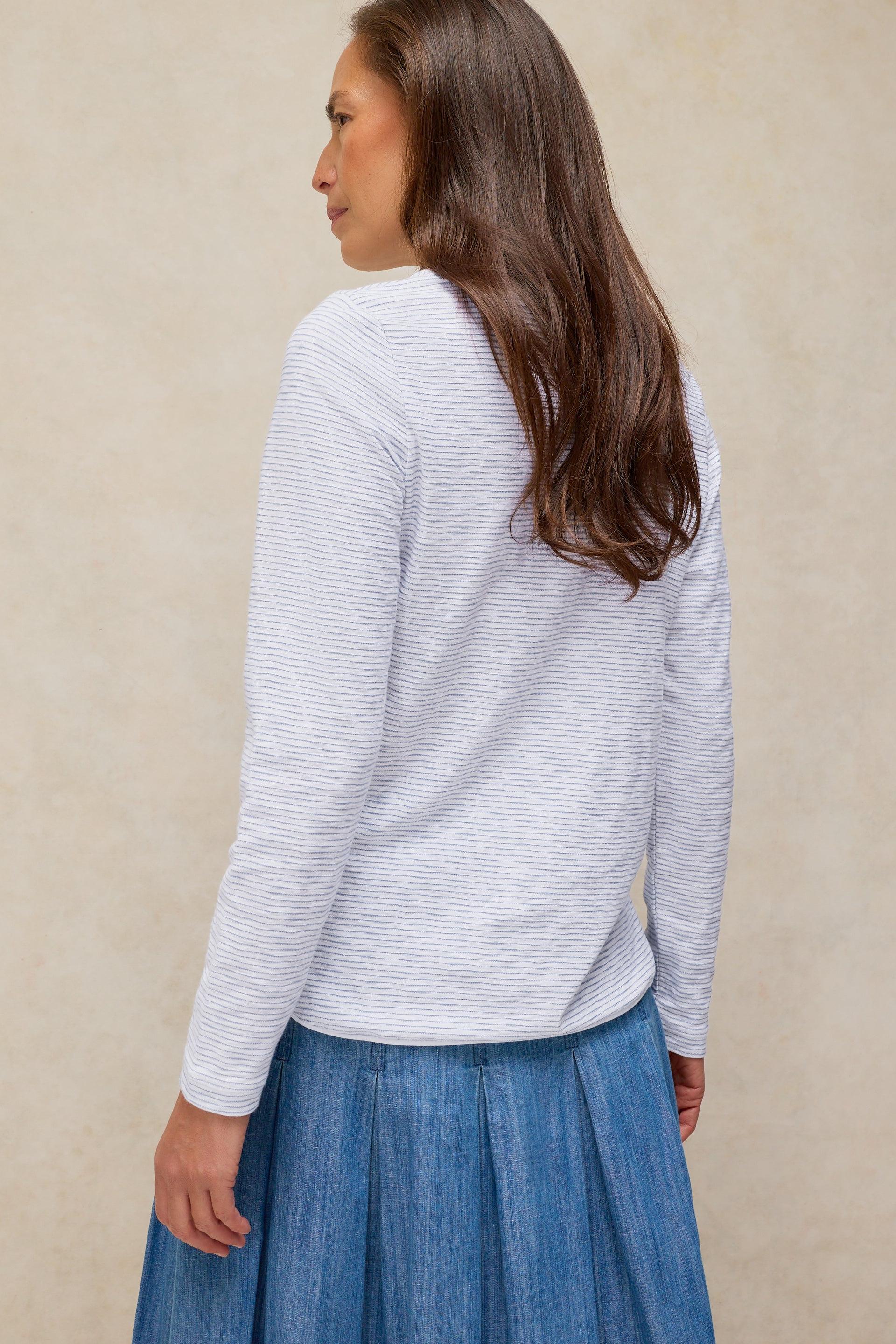 A woman seen from the back wearing a long-sleeved striped cotton top and a blue pleated skirt.
