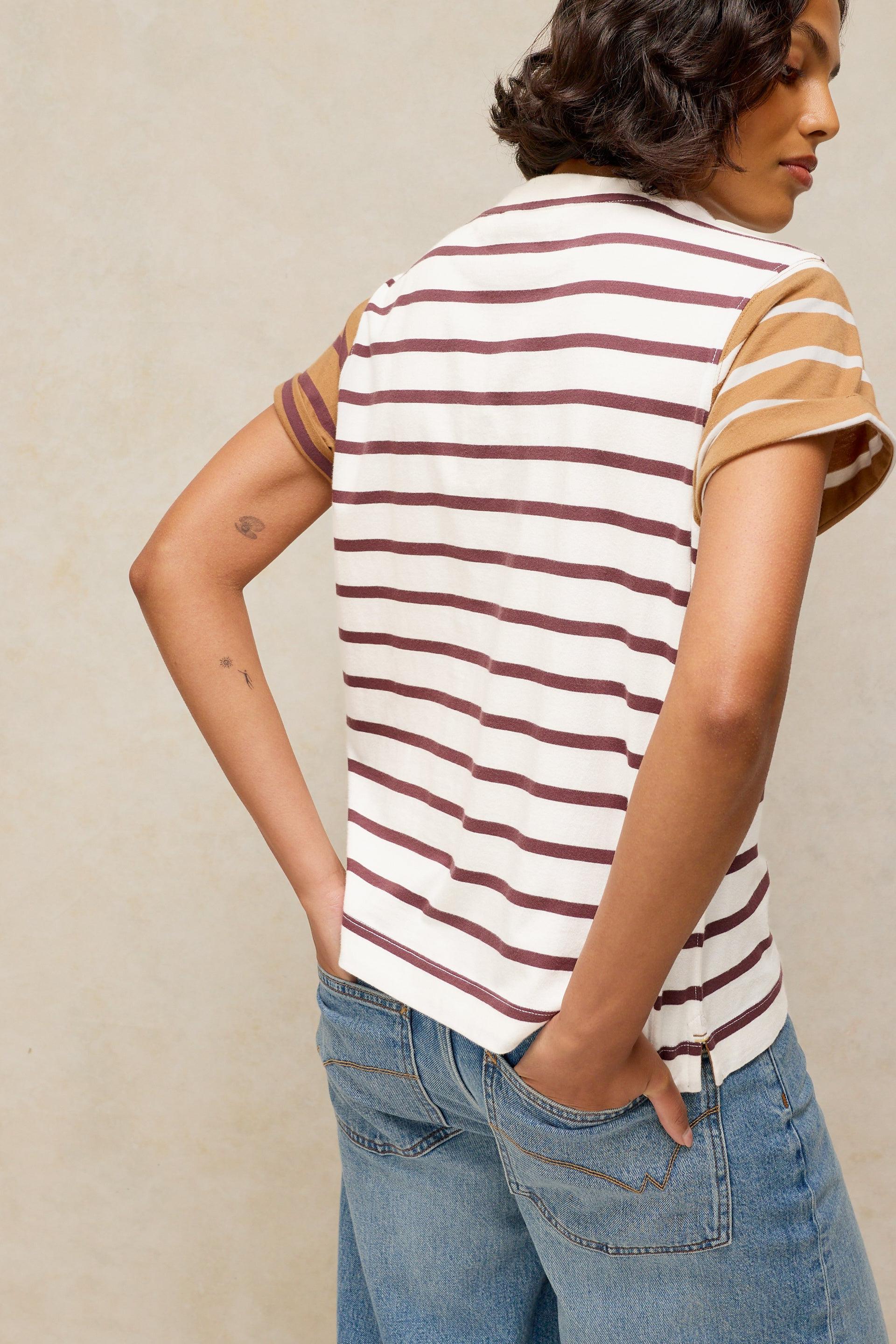A model shown from the back wearing a sleeveless striped top with white, beige, and dark brown horizontal stripes, paired with blue jeans, against a neutral beige background.