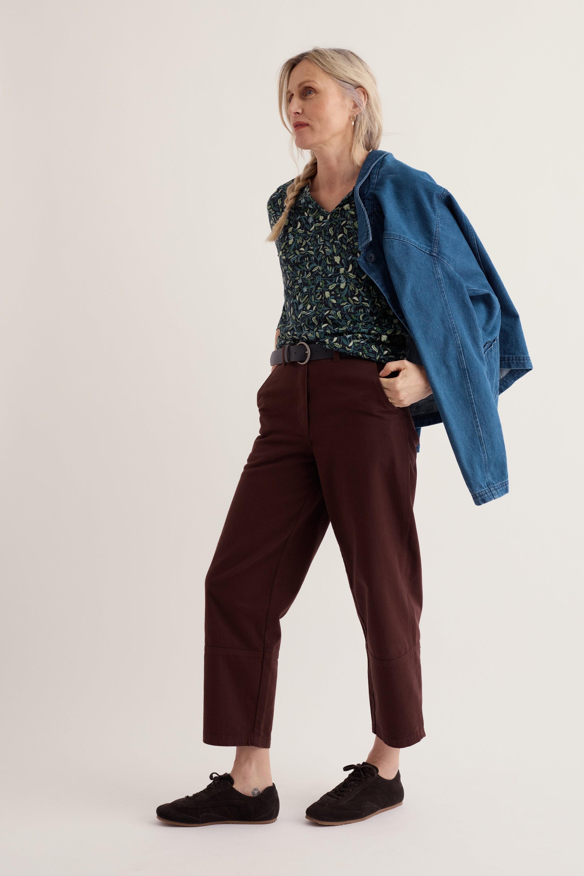 A woman modeling a floral organic cotton top, brown trousers, and a blue denim jacket over her shoulder.