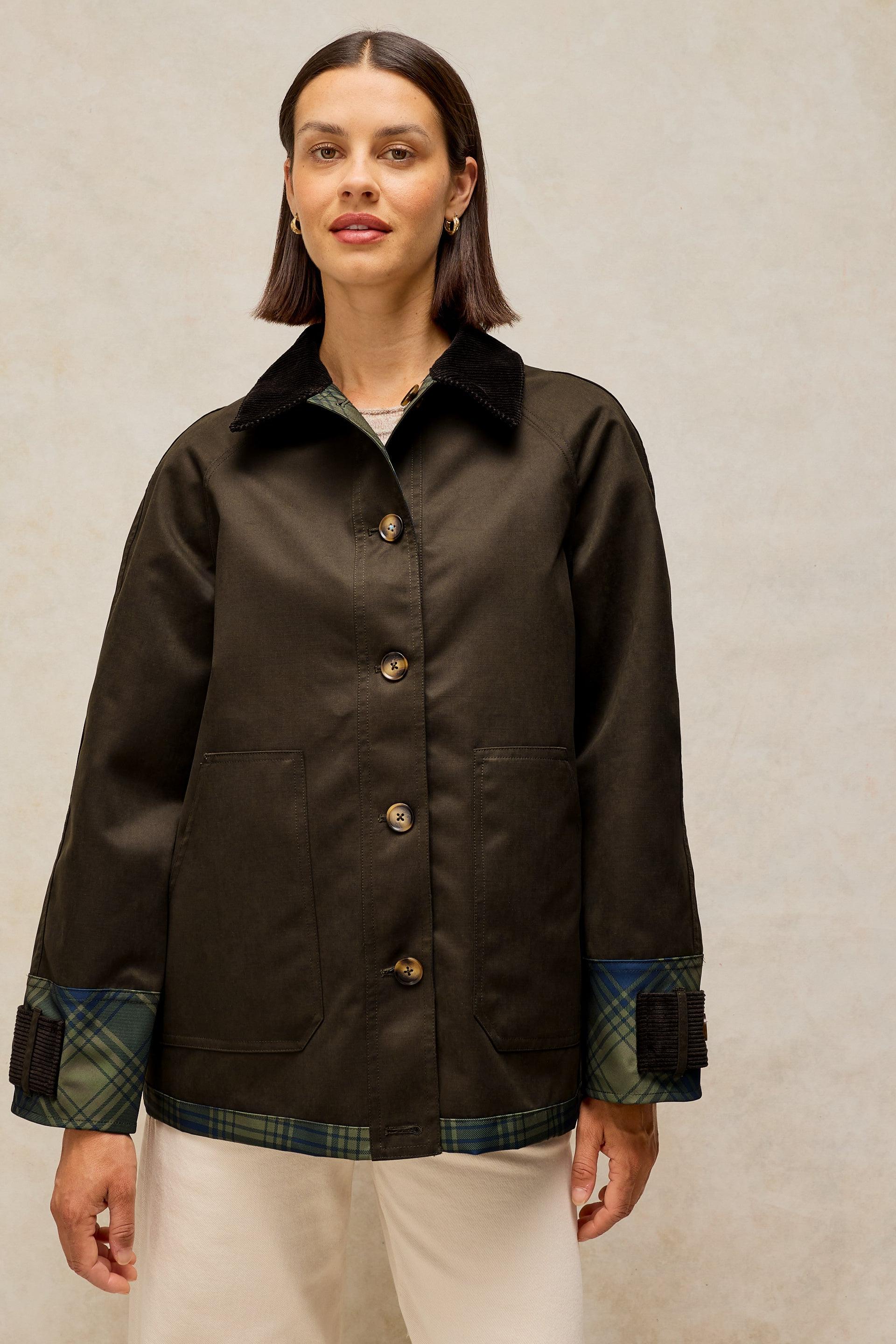 A woman modeling a dark brown waxed cotton jacket with green and blue plaid trim on the cuffs and hem, paired with cream trousers against a textured beige background.