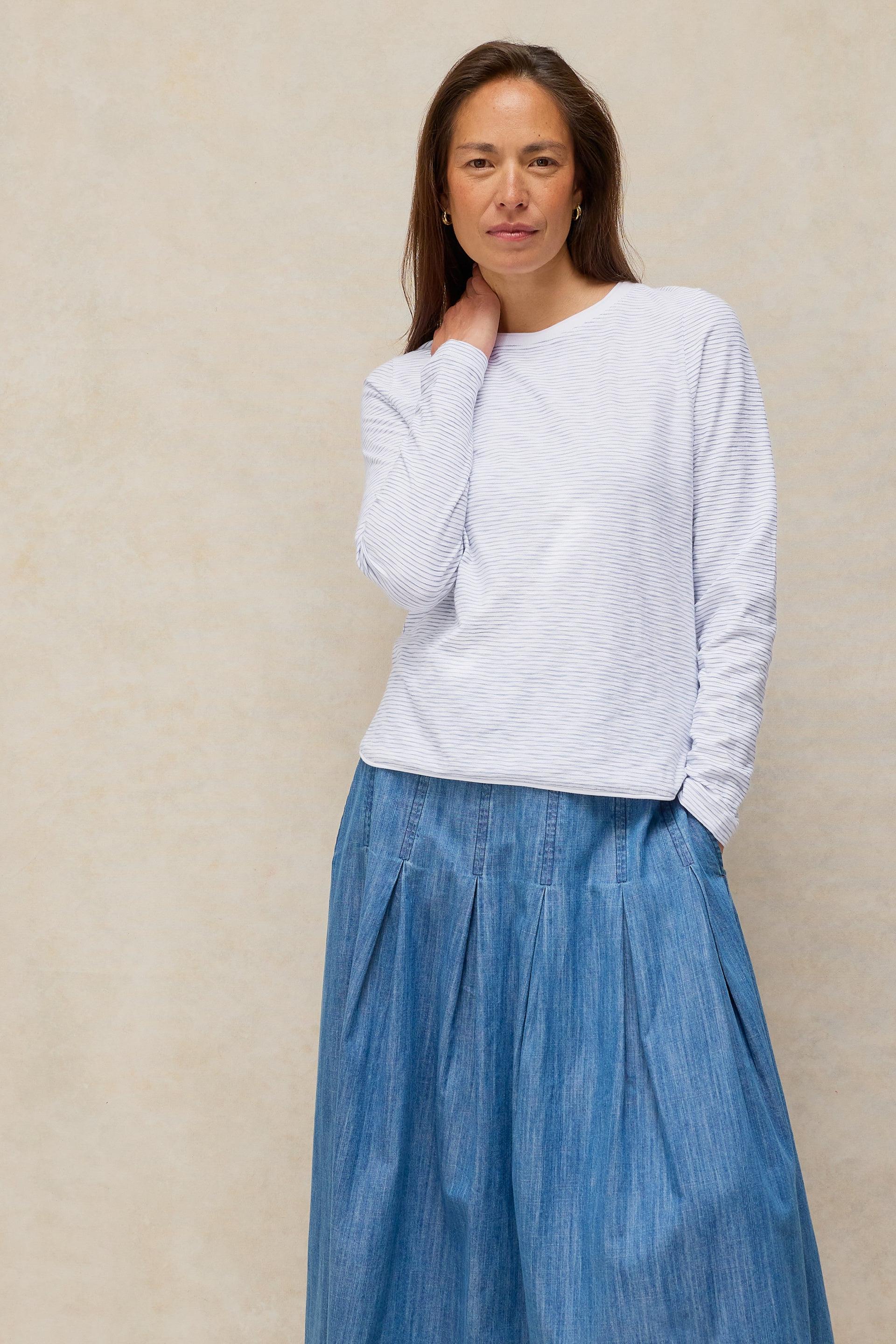 Woman wearing a long-sleeved white cotton striped top and a blue denim skirt standing against a beige wall.