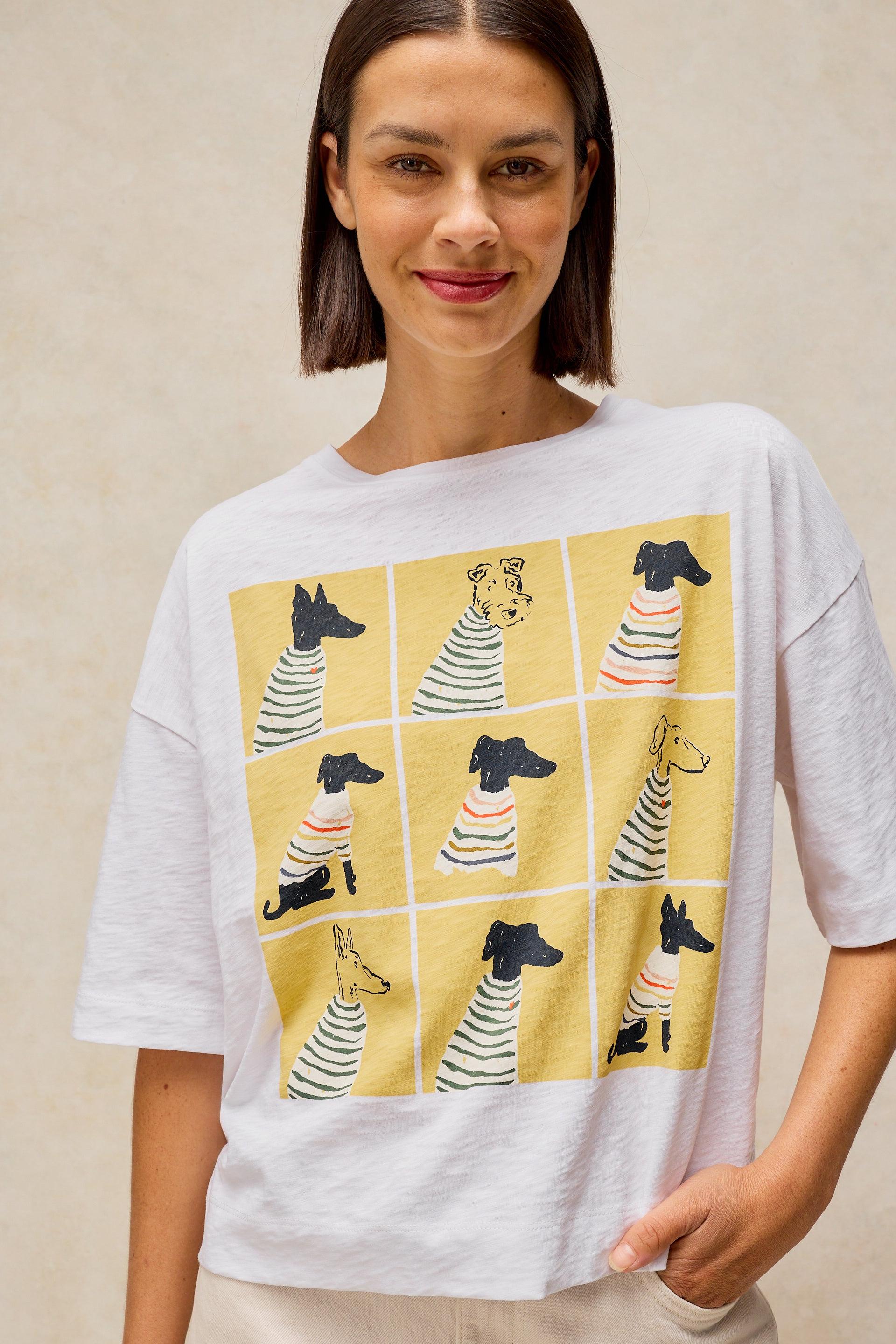 A woman wearing a white oversized cotton t-shirt with a yellow graphic print of cartoon dogs in striped shirts, posed against a neutral beige background.