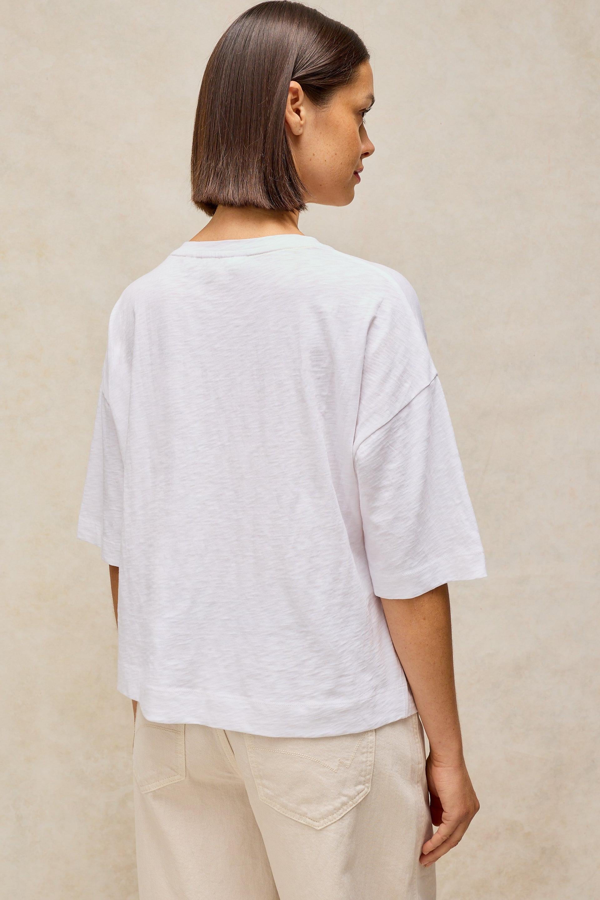 A woman with a shoulder-length brown bob is shown from the back wearing a white short-sleeve crewneck t-shirt and beige pants, set against a textured beige wall.