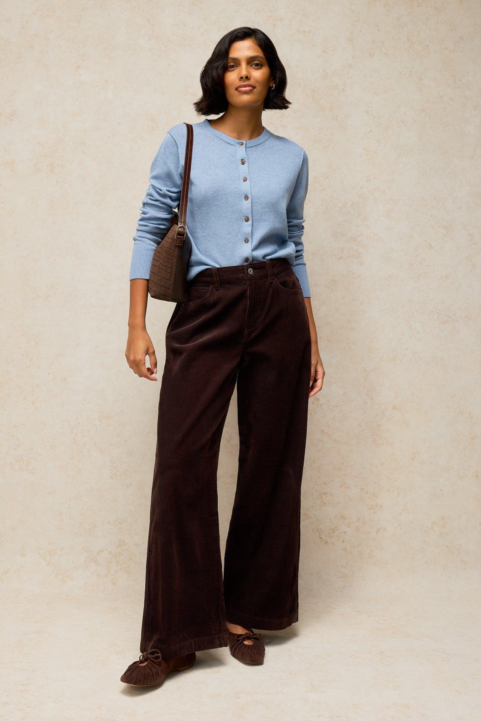 A woman modeling brown cord trousers from White Stuff with a blue buttoned cardigan.