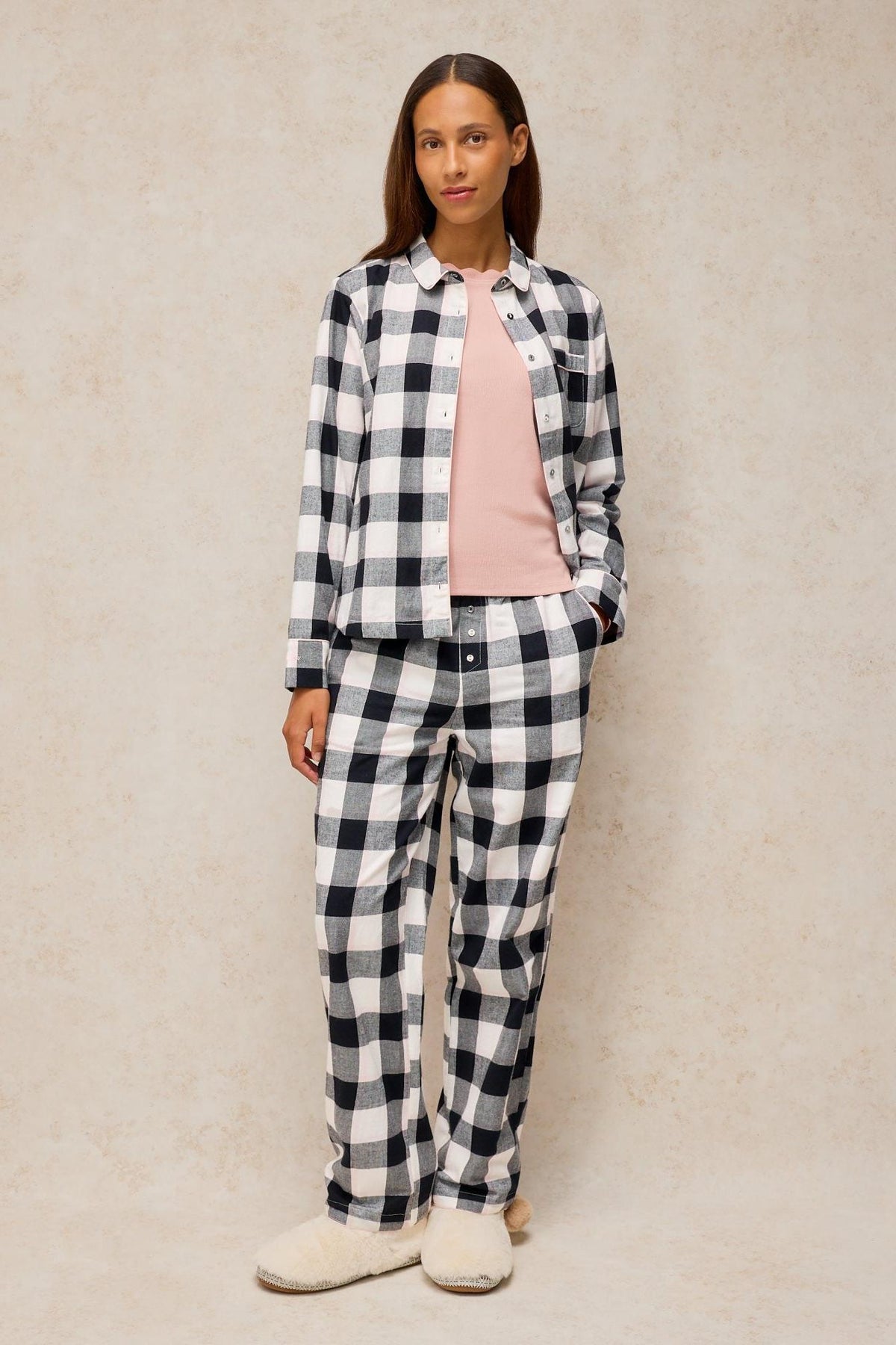 Woman standing wearing a black and white check organic cotton pyjama set and cosy slippers.