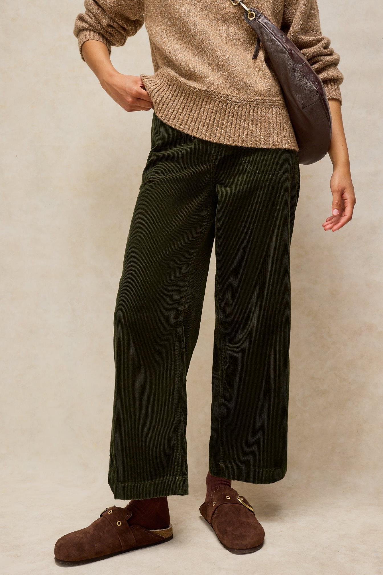 Woman posing in dark green White Stuff cord trousers with a beige knit sweater and brown suede mules.