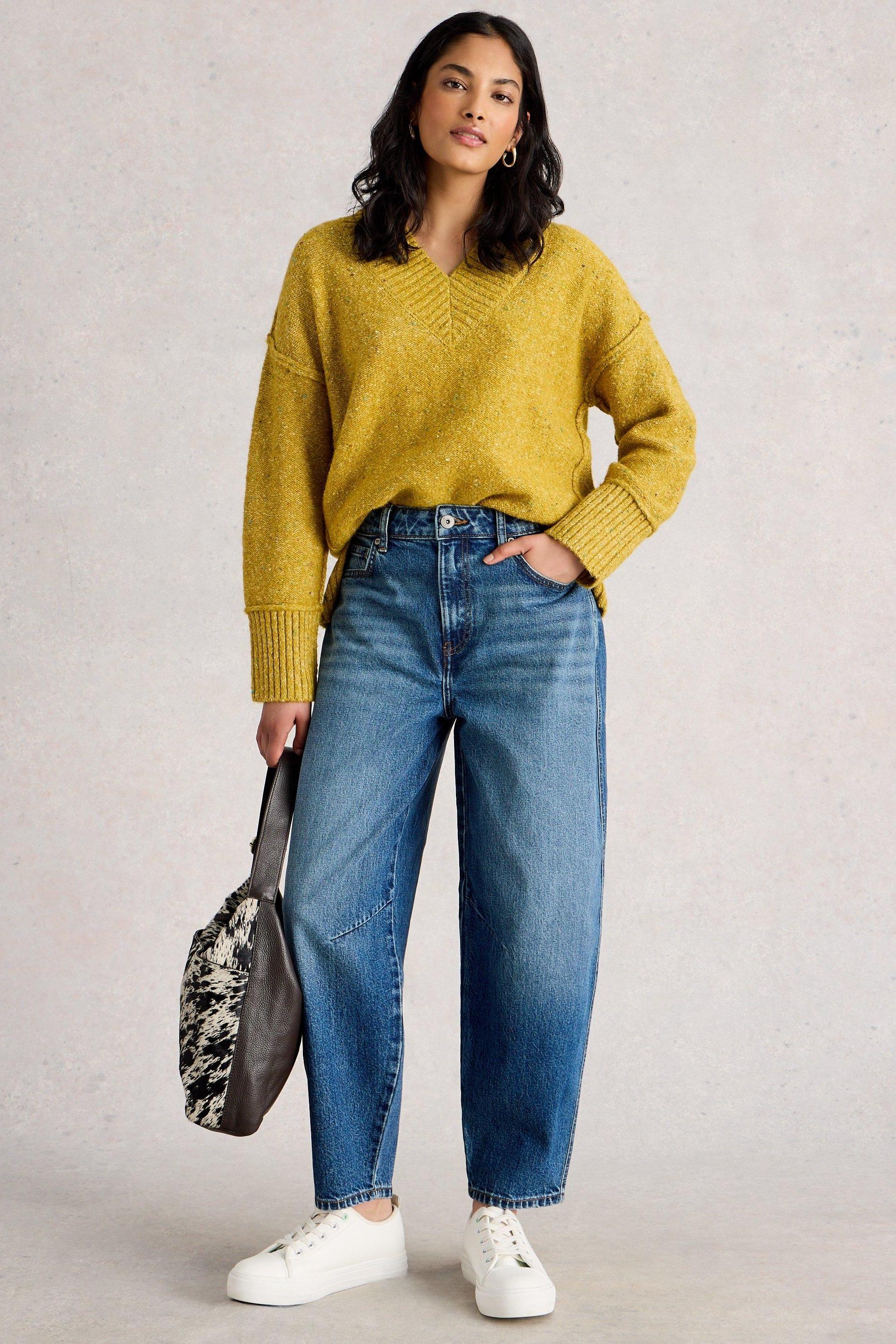 A woman modeling White Stuff mid denim barrel leg jeans with a yellow sweater and white sneakers.