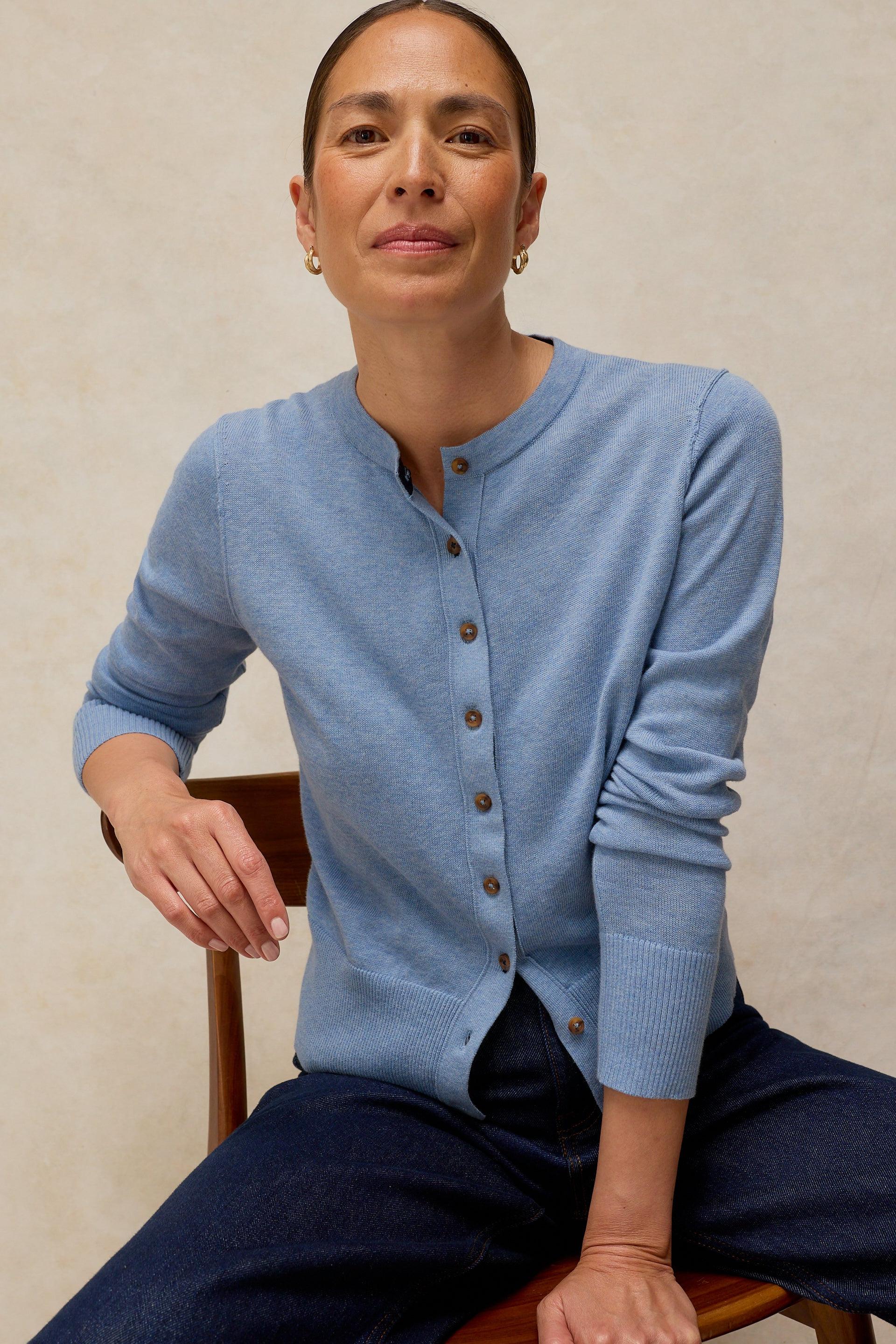 A woman wearing a soft blue White Stuff organic cotton button-up cardigan sitting on a wooden chair