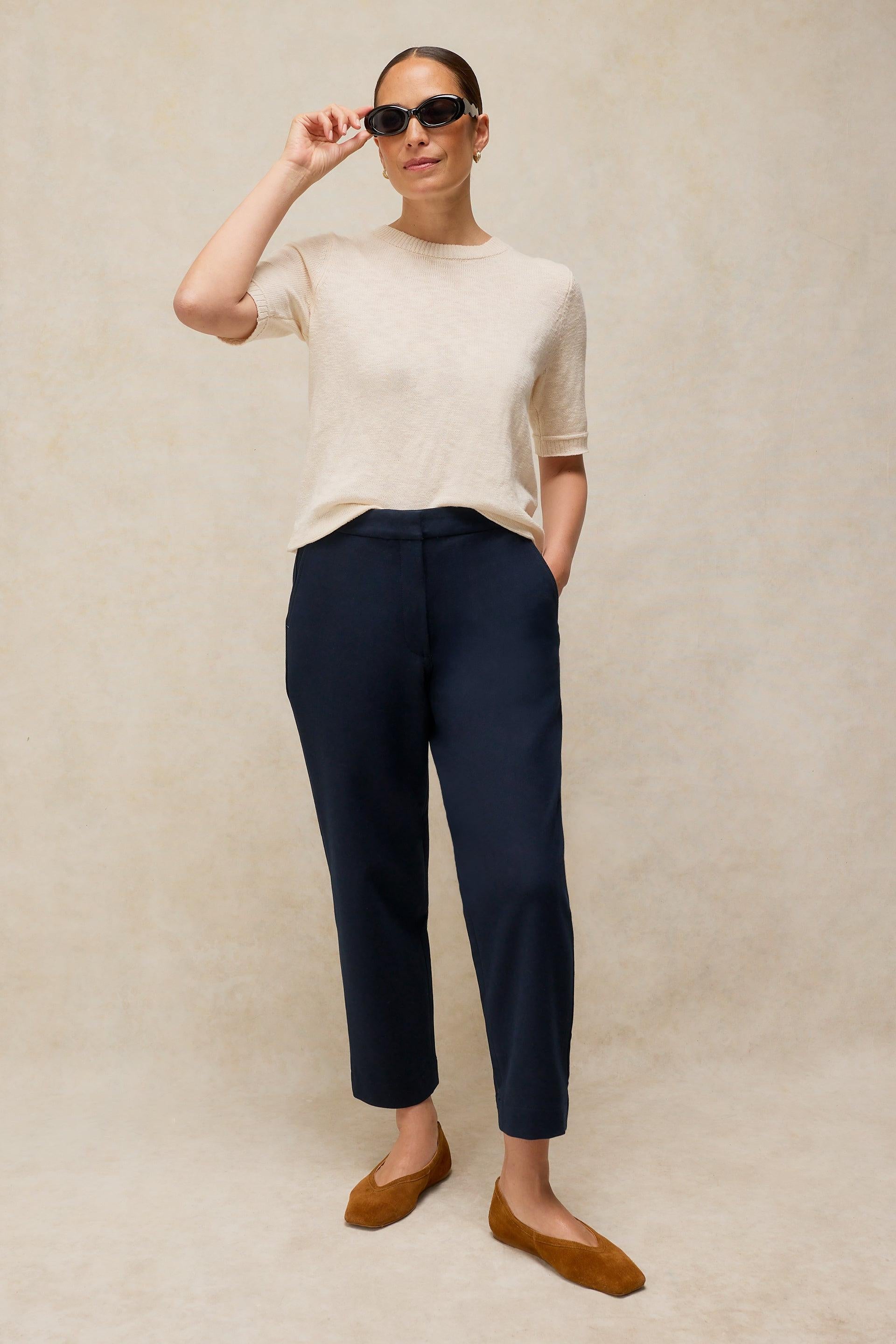 A woman wearing a white organic cotton t-shirt, navy trousers, and sunglasses against a neutral studio backdrop