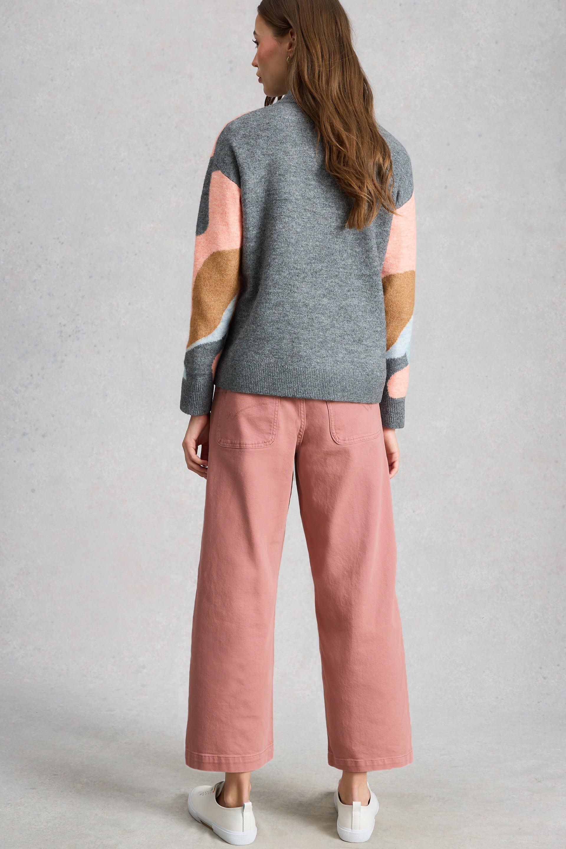 Woman standing with back turned wearing a grey patterned sweater and pink organic cotton trousers.
