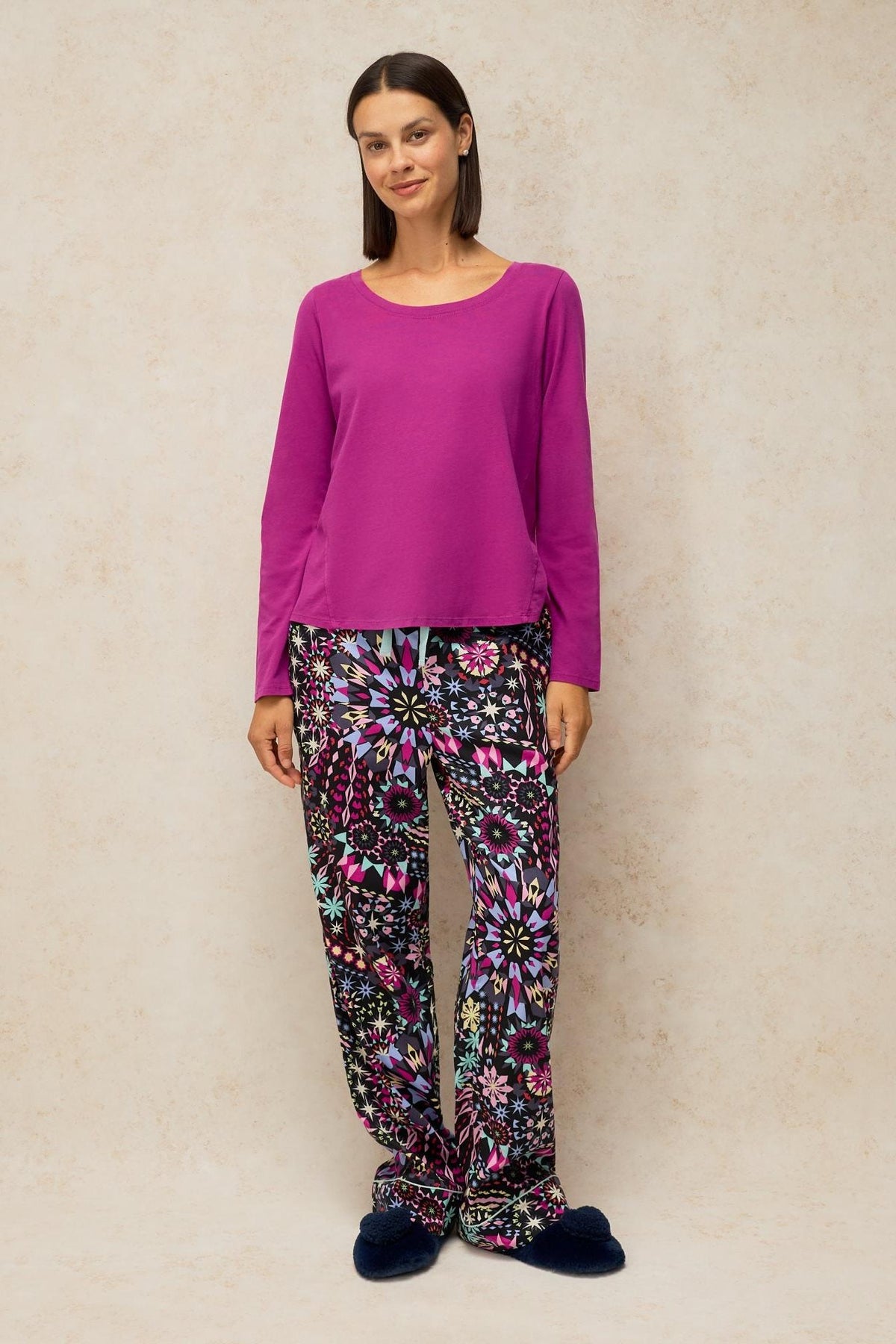 A woman wearing a pink long-sleeved organic cotton pyjama top and patterned trousers against a neutral background.