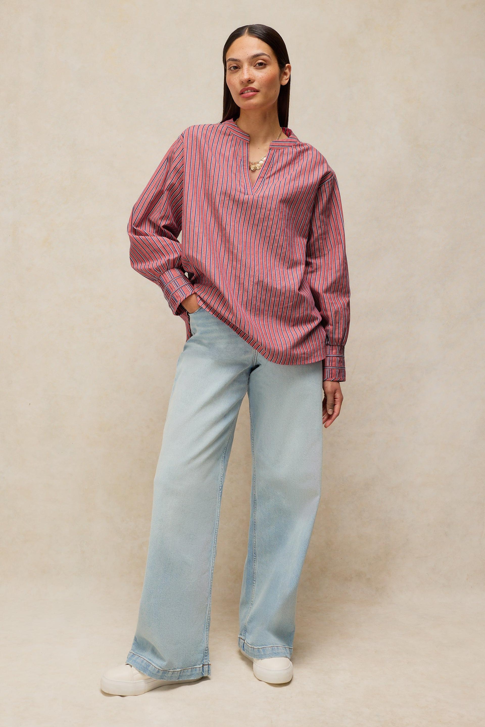 A woman modeling a loose-fitting, long-sleeve button-up shirt with vertical pink and white stripes, paired with light-wash wide-leg jeans and white sneakers, against a neutral beige textured studio backdrop.