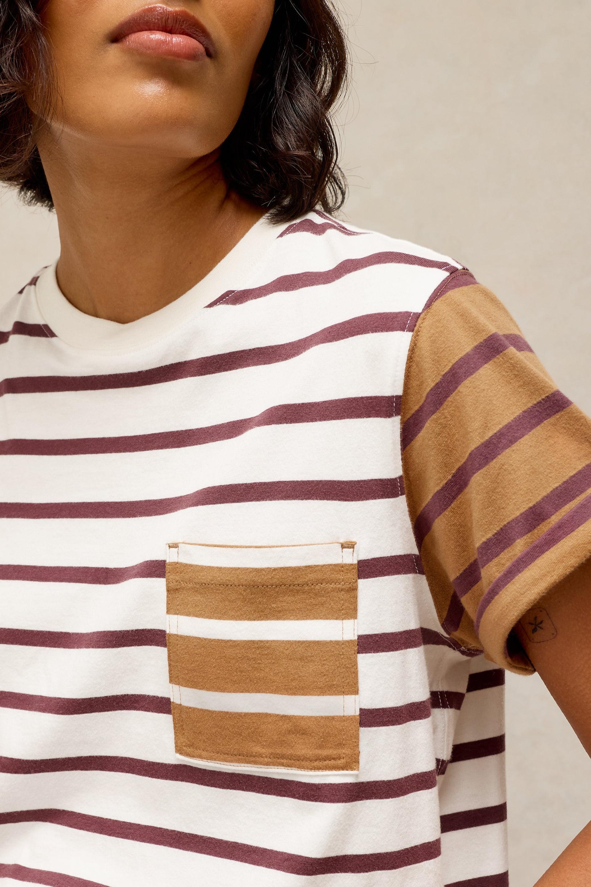 Close-up of a person wearing a short-sleeve crew-neck T-shirt with horizontal burgundy stripes on an off-white base, plus a tan chest pocket and a solid tan sleeve.