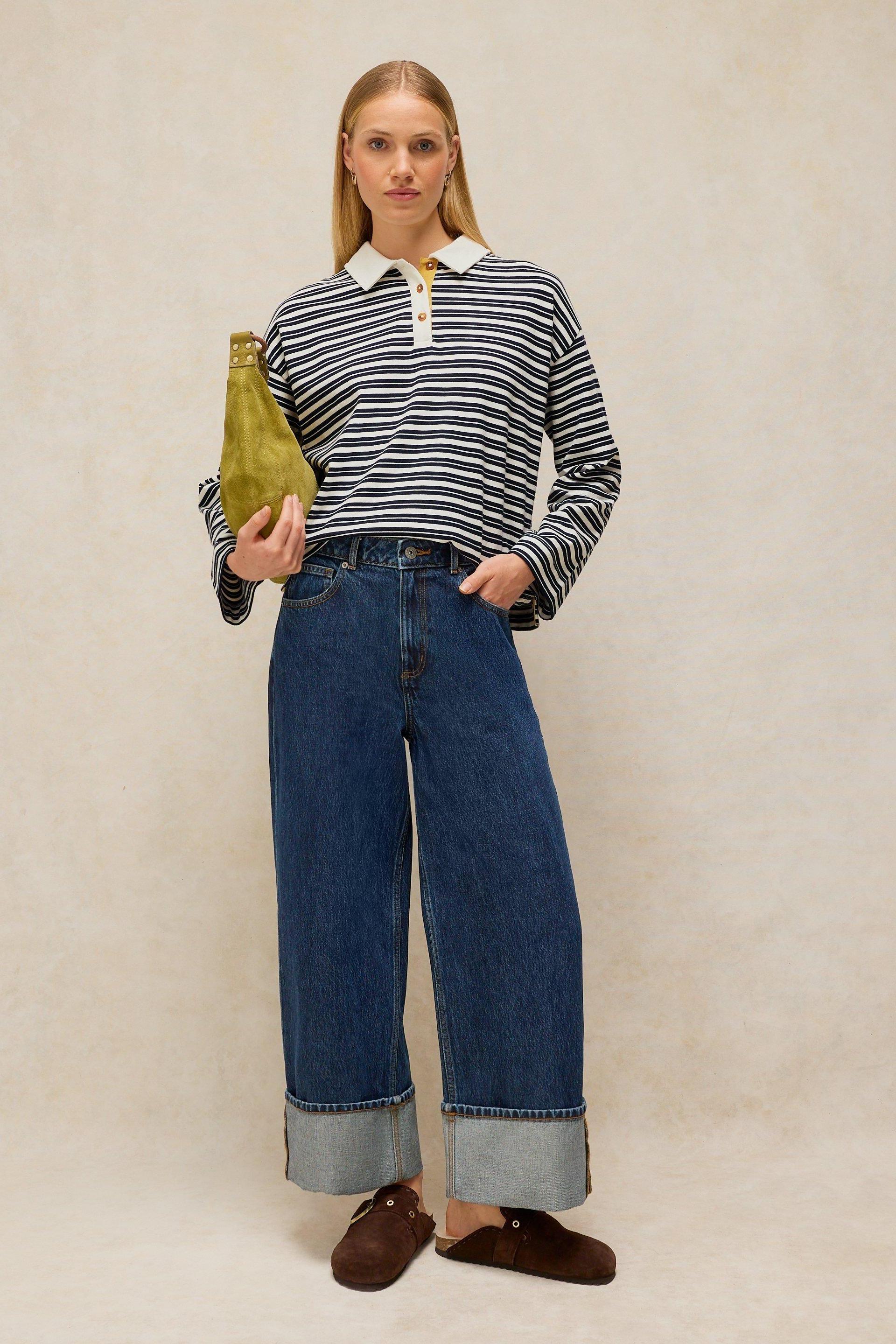 A woman posing in a White Stuff striped women's rugby top with wide-leg jeans and an olive green handbag.