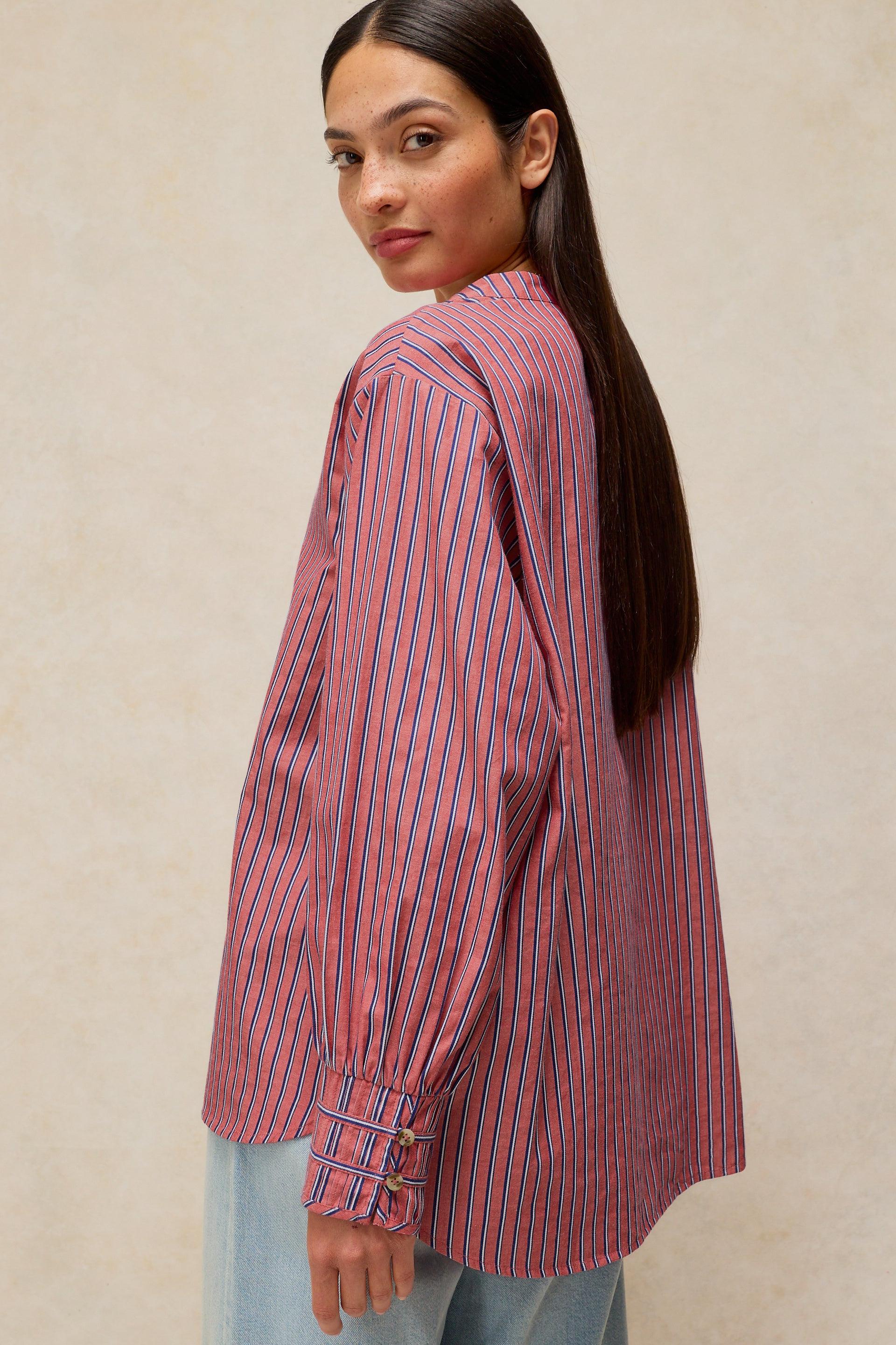 A woman wearing a pink and blue vertically striped button-up shirt, shown from a side-back angle against a neutral beige textured background.