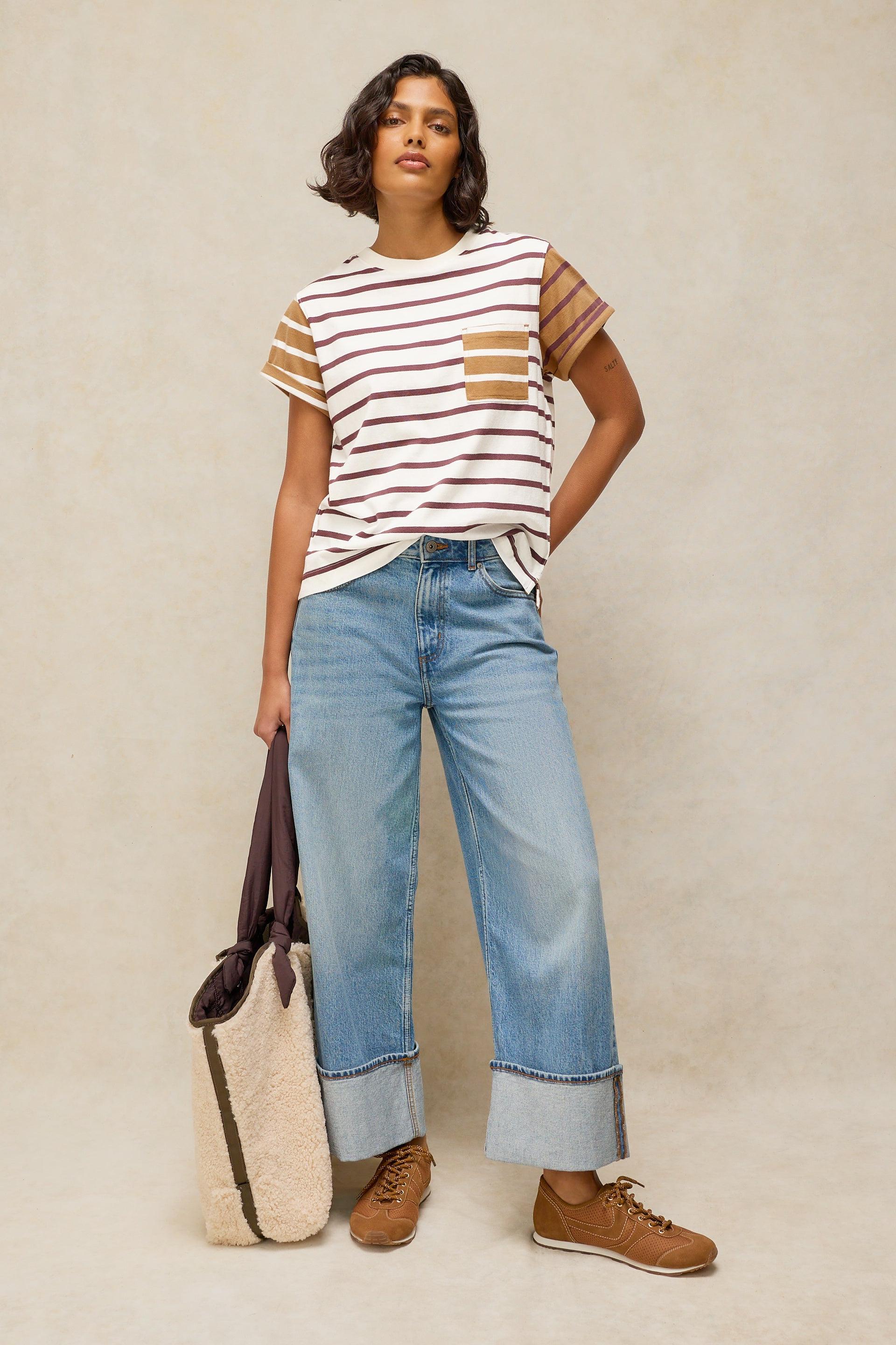 A woman stands against a plain beige studio background wearing a cream short-sleeve striped T-shirt with tan patch pockets, light blue wide-leg cuffed jeans, and brown lace-up shoes. She holds a large cream tote bag with dark trim.
