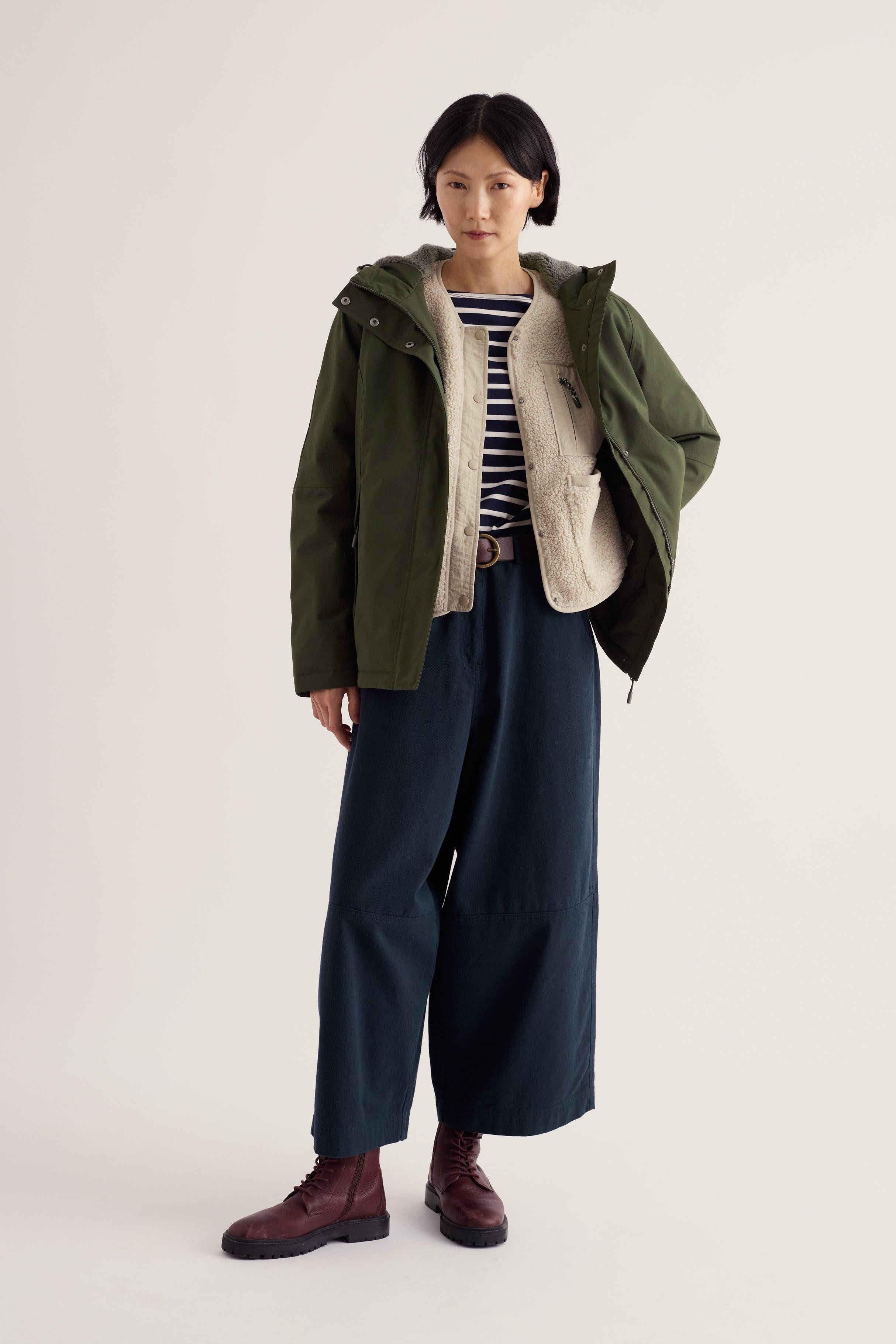 A woman wearing a woodland green Seasalt Cornwall waterproof coat with a sherpa-lined vest and striped top.