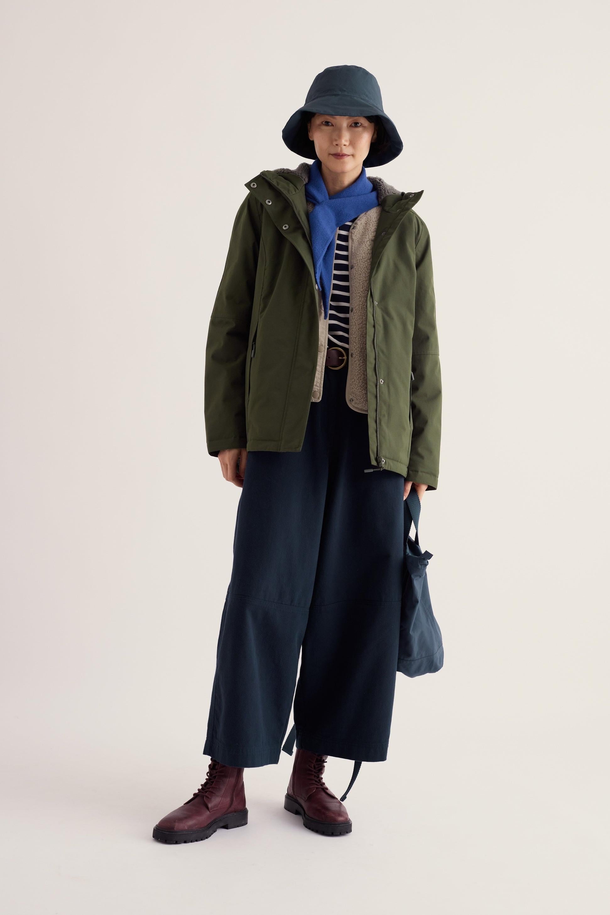 A woman posing in a woodland green Seasalt Cornwall waterproof coat, bucket hat, and wide-leg trousers.