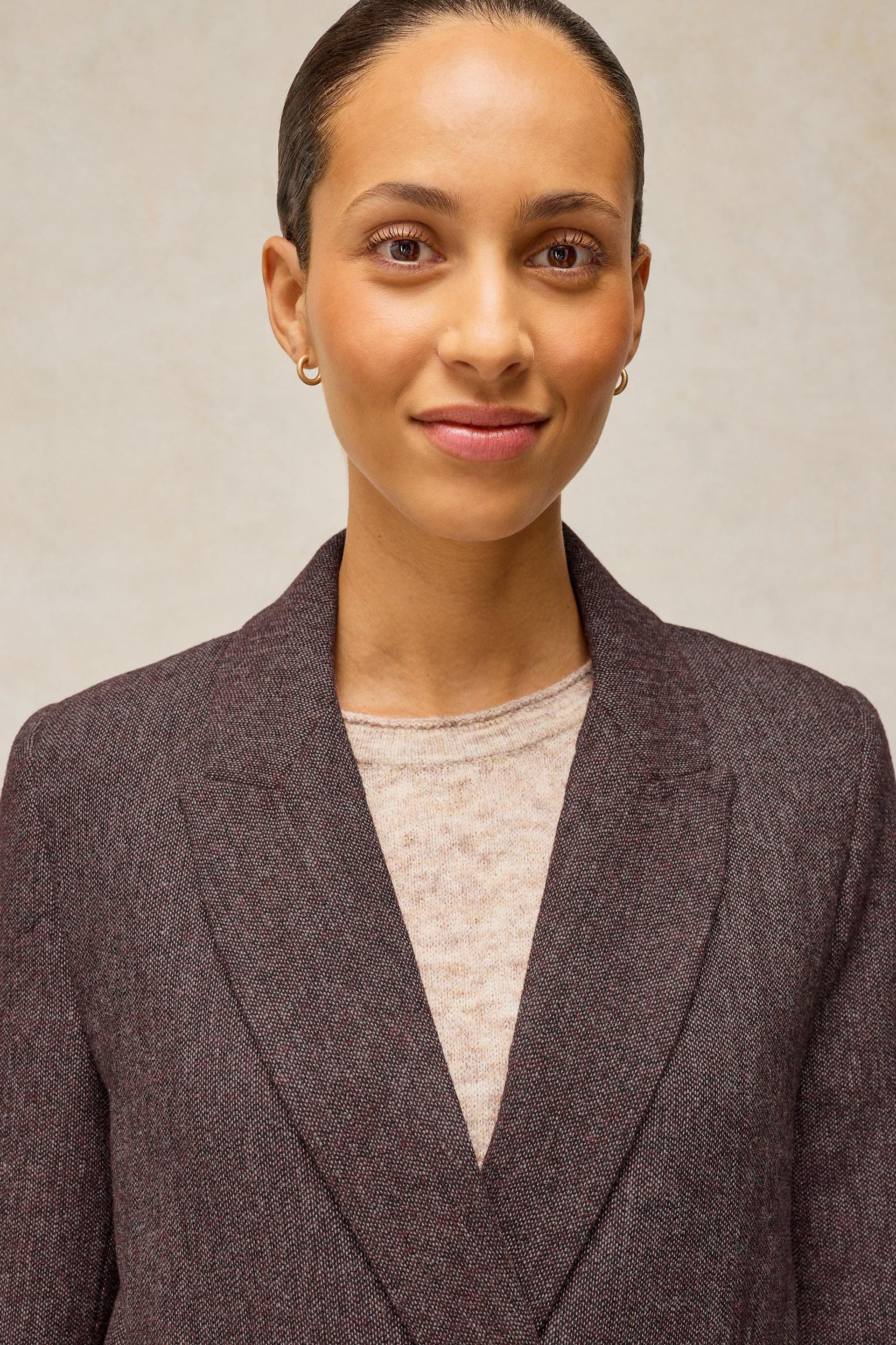 A woman wearing a dark brown textured blazer over a light beige knit top, photographed against a neutral textured beige background.