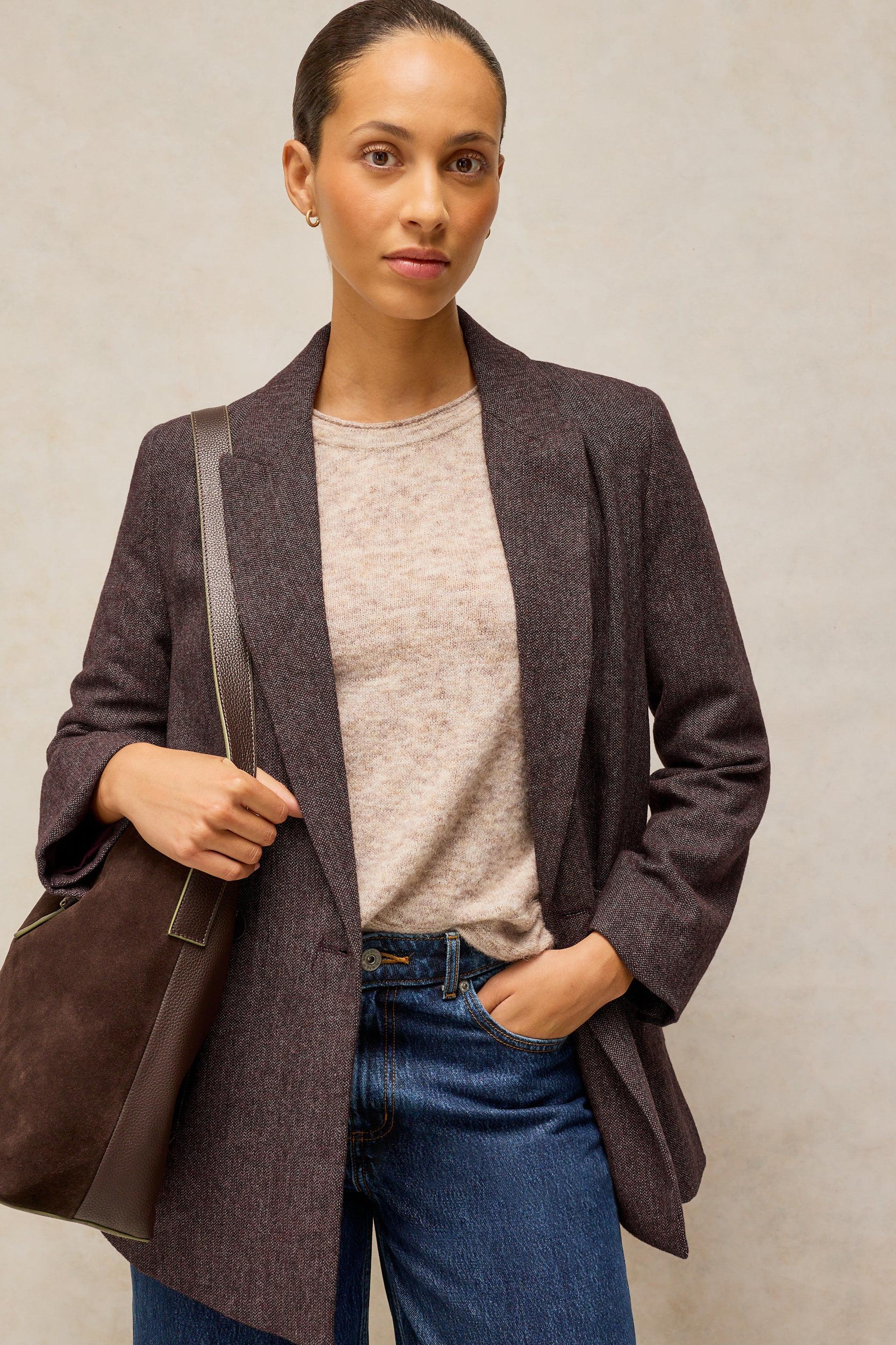 A woman wearing a dark plum open-front blazer over a light beige crewneck sweater and blue jeans, carrying a dark brown leather shoulder bag against a textured light gray background.