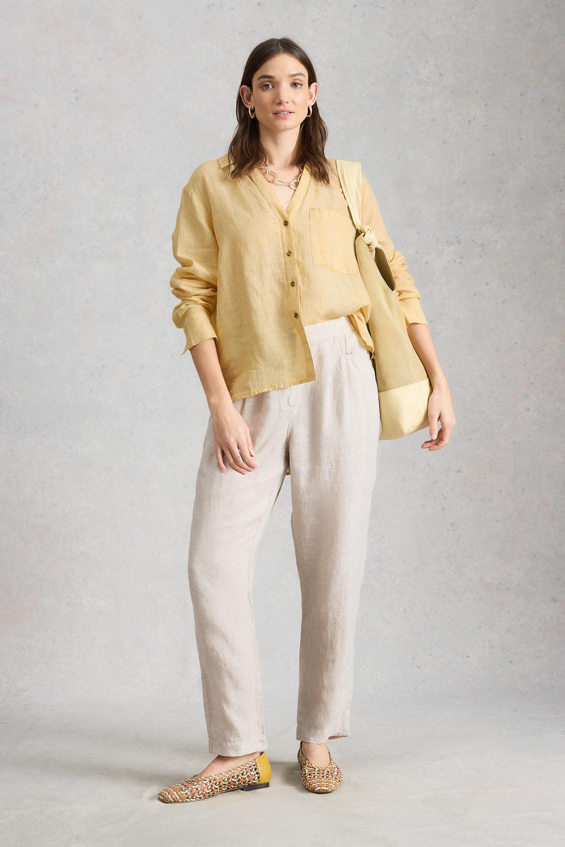 Woman wearing a yellow striped linen shirt and beige linen trousers, standing against a neutral studio background.