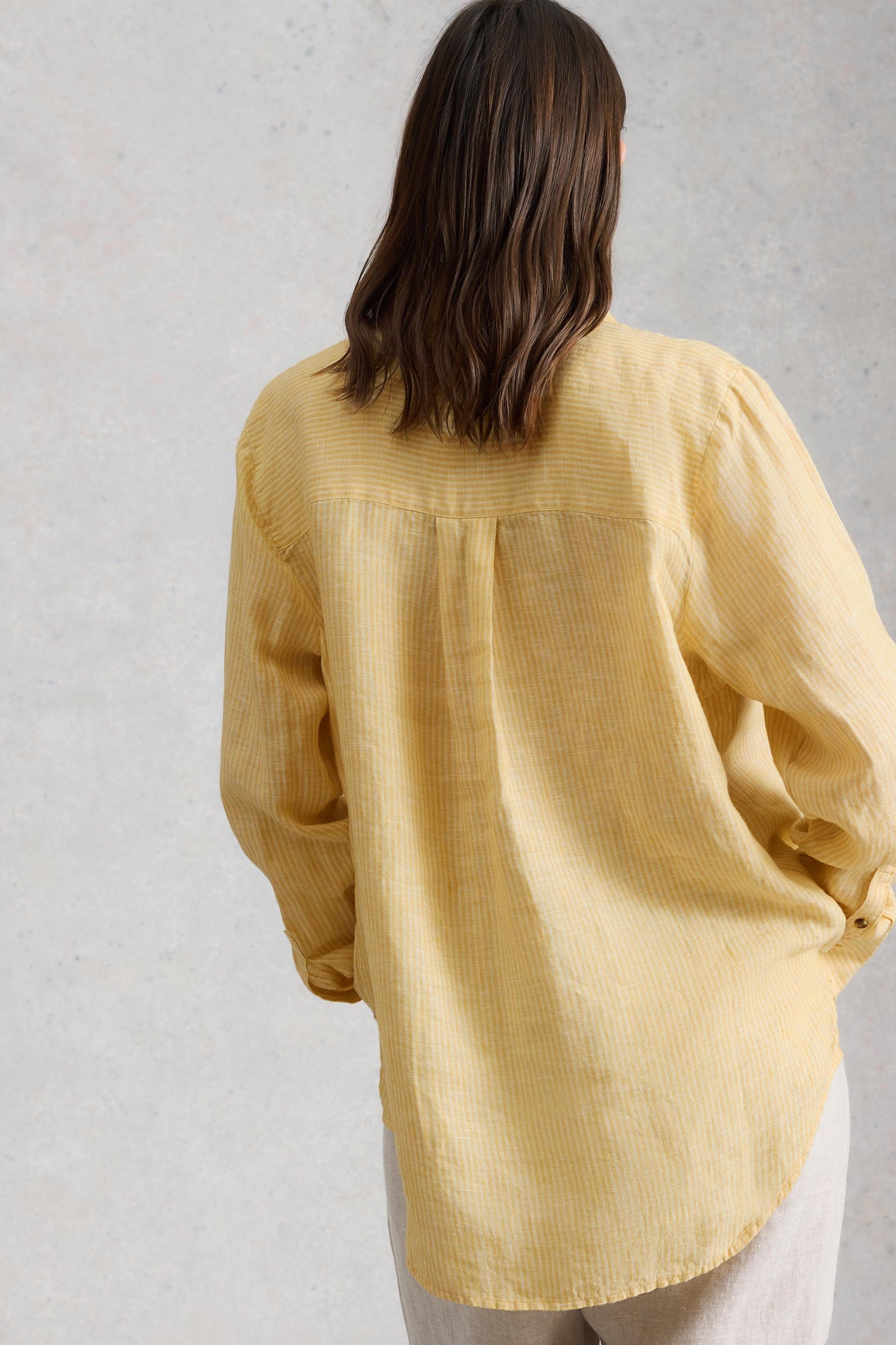 A woman seen from the back wearing a yellow striped long-sleeve linen shirt against a grey textured background.