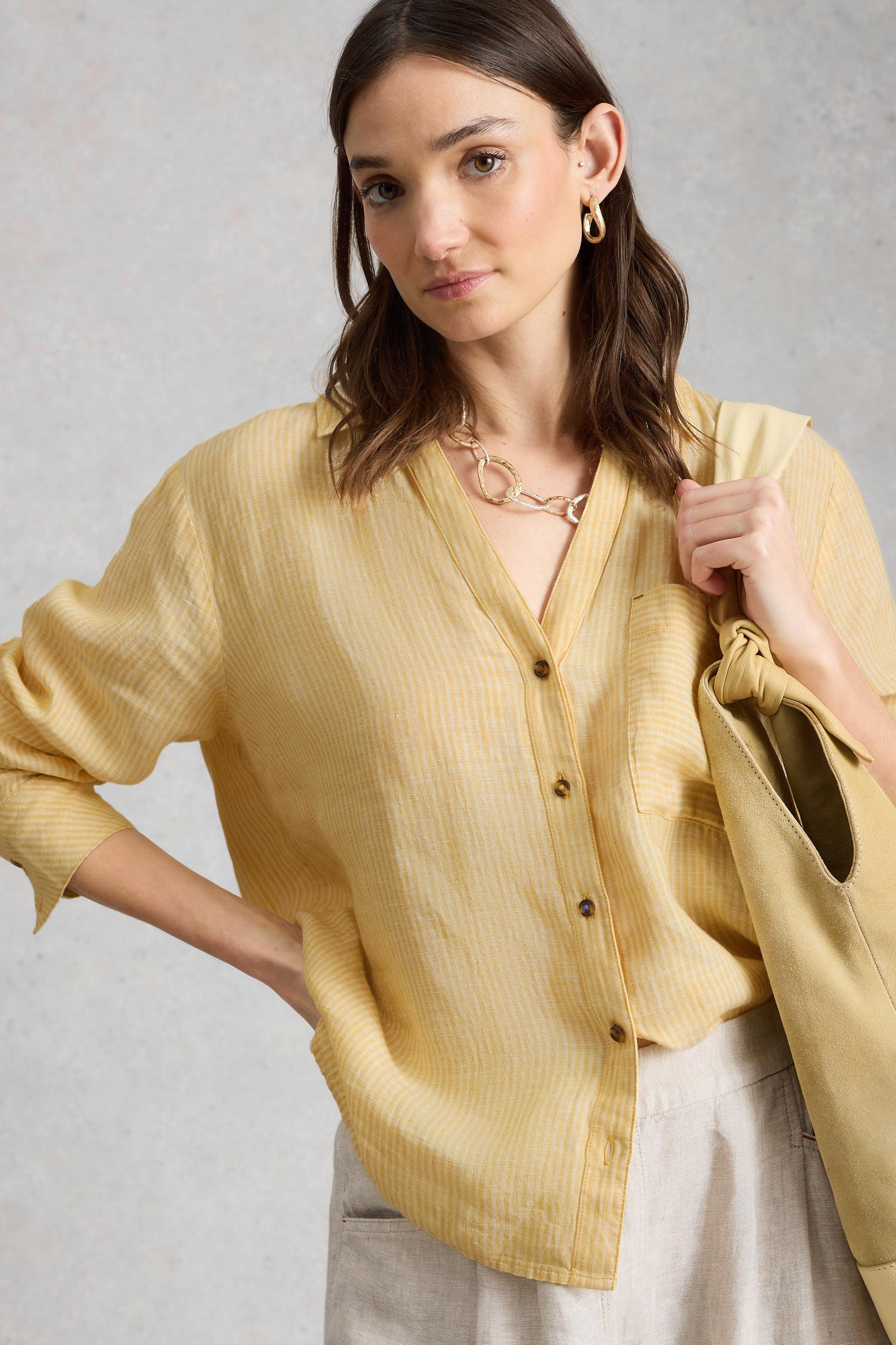 Woman wearing a yellow striped linen shirt and carrying a suede shoulder bag against a neutral studio background.