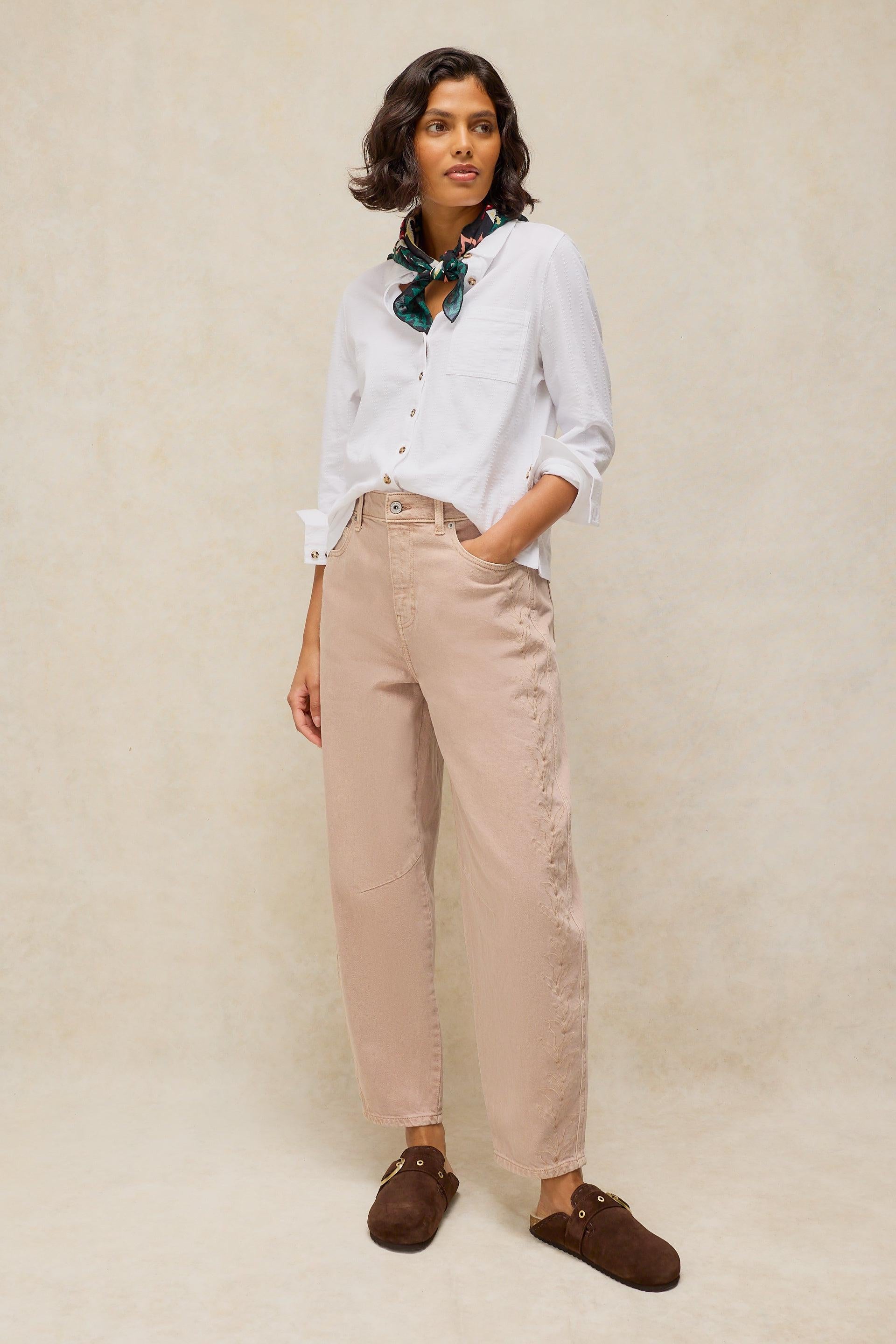 A woman wearing a white organic cotton shirt and tan barrel jeans against a neutral background.