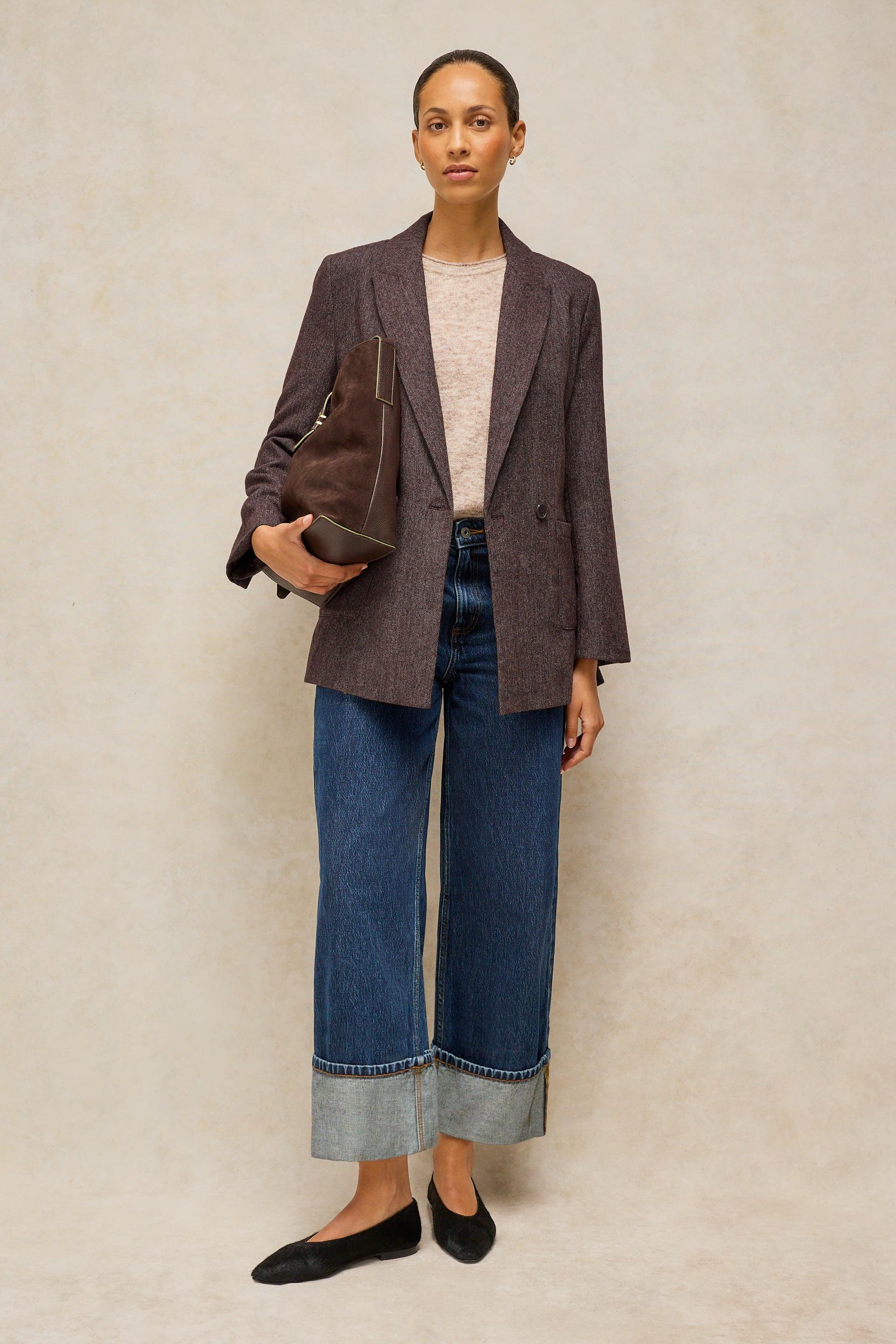 A model stands against a textured beige studio backdrop wearing a dark brown textured blazer, a light beige knit top, wide-leg cropped blue jeans with rolled cuffs, black flats, and holding a dark brown leather tote bag.