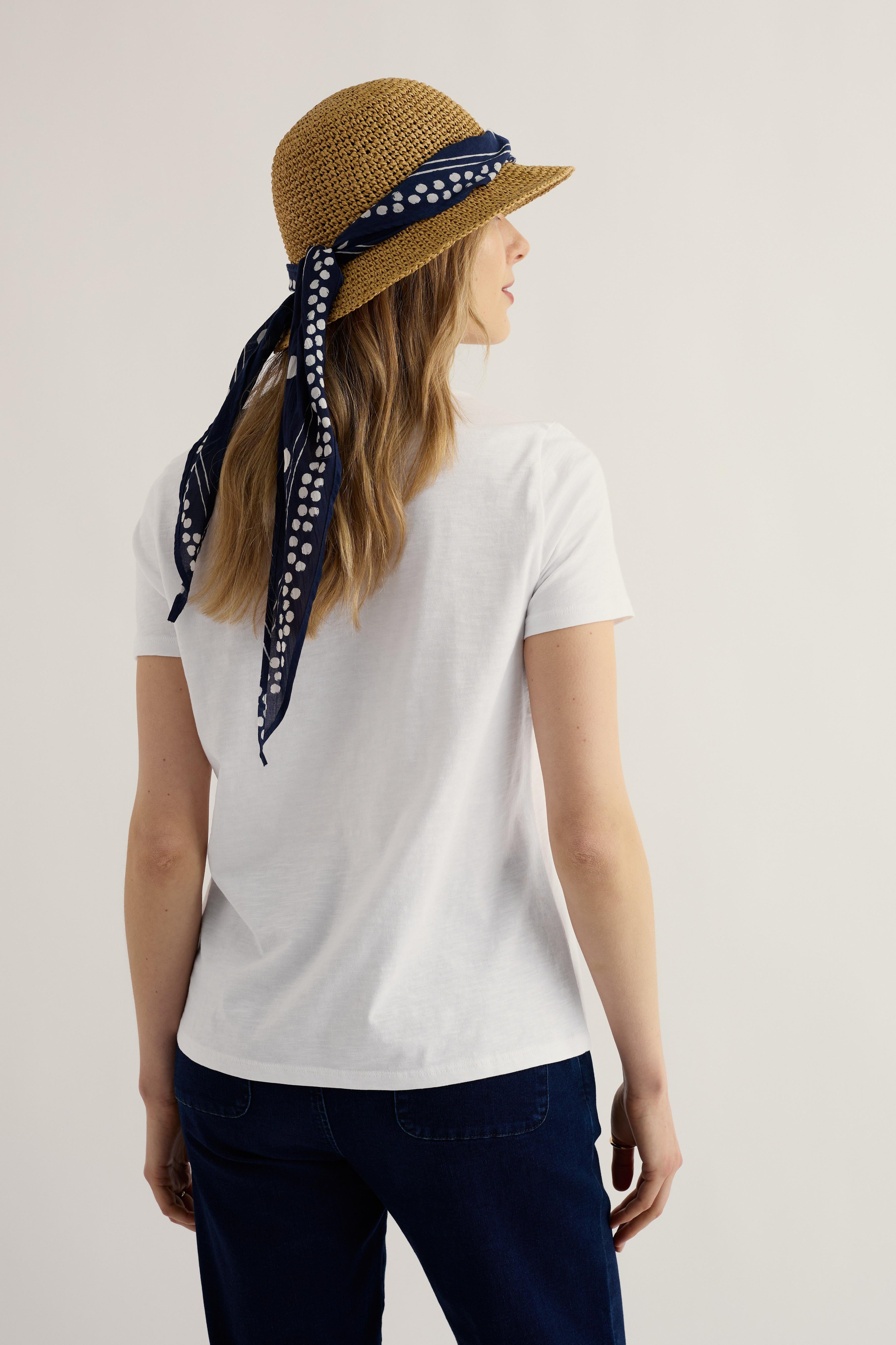 Woman wearing a white organic cotton t-shirt, navy pants, and a straw hat with a blue bandana, standing against a plain background.