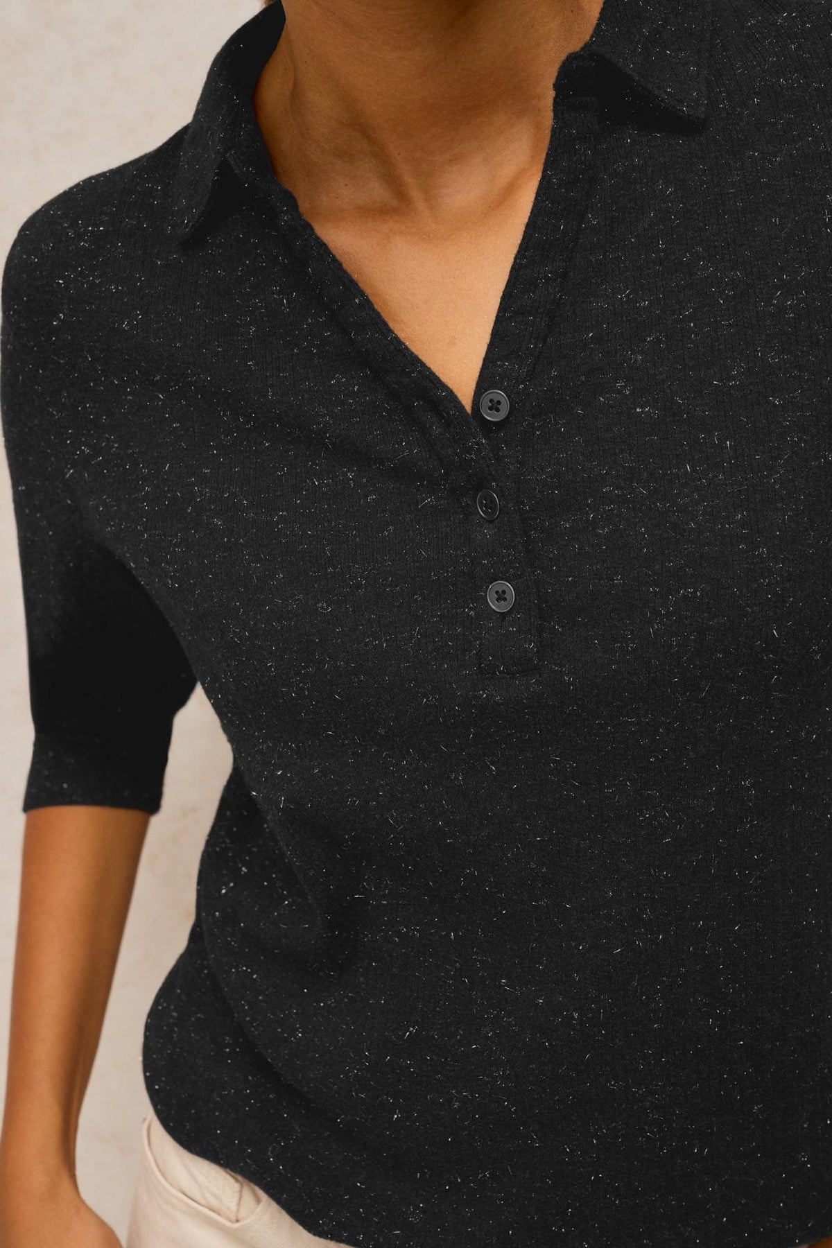 A woman wearing a black ribbed cotton polo top with subtle sparkle detailing.