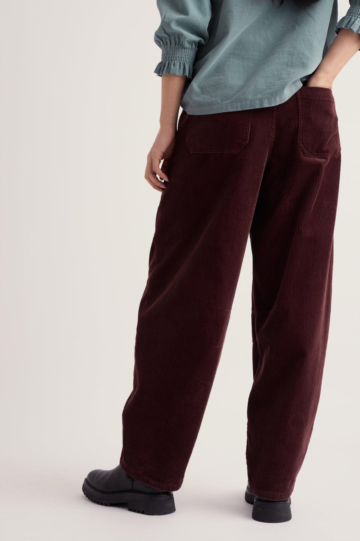 A person wearing brown barrel leg cord trousers and a blue top against a light background.