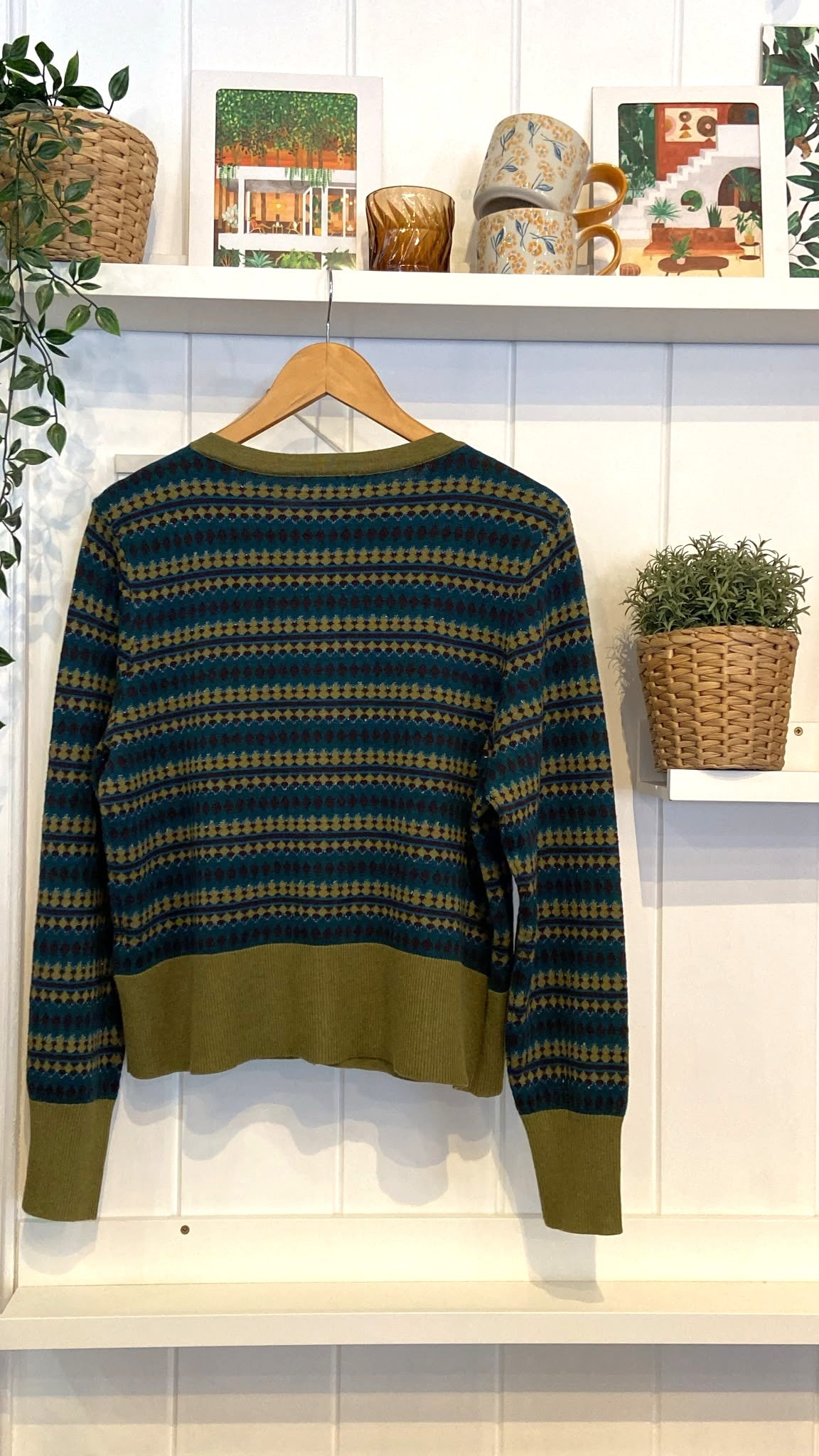 A women's Fair Isle pattern cardigan with green trim hanging on a wooden hanger against a white wall.