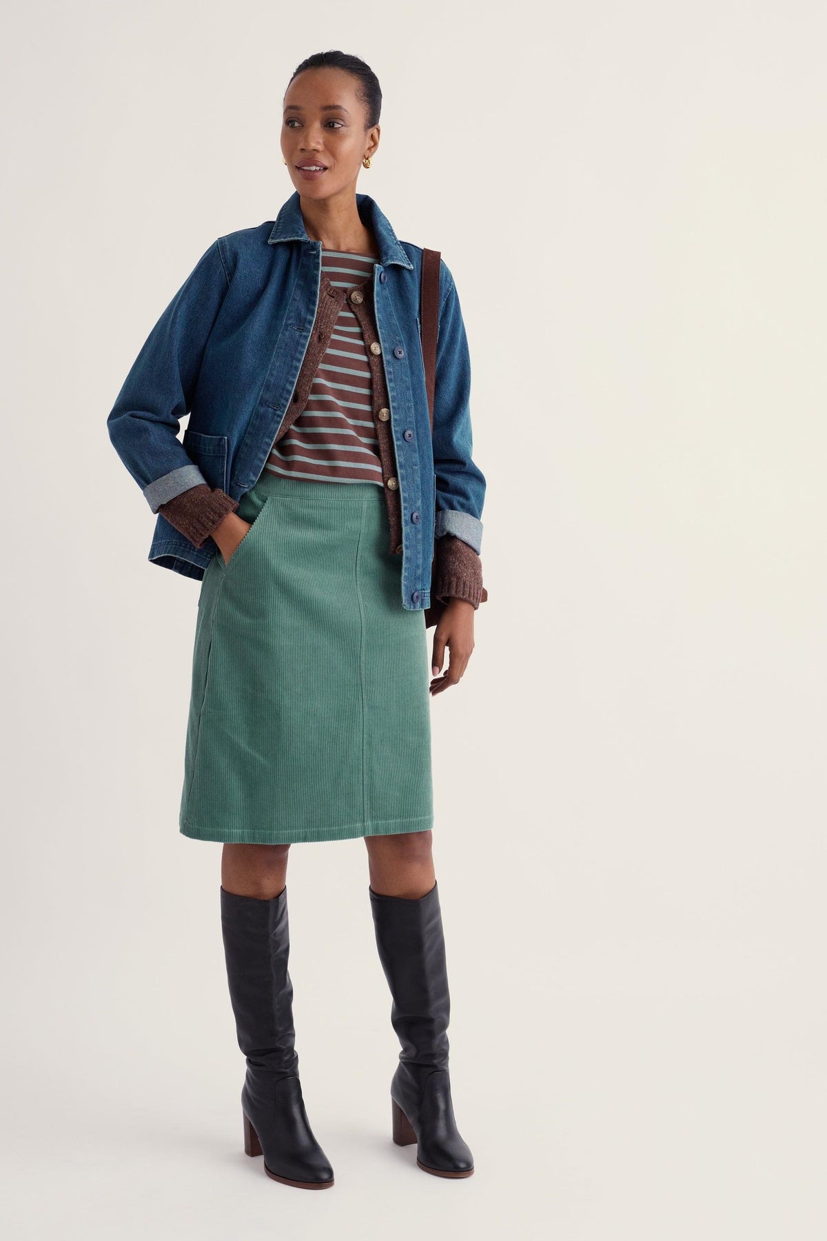 A woman wearing a knee-length green corduroy Seasalt skirt styled with a denim jacket and black boots.