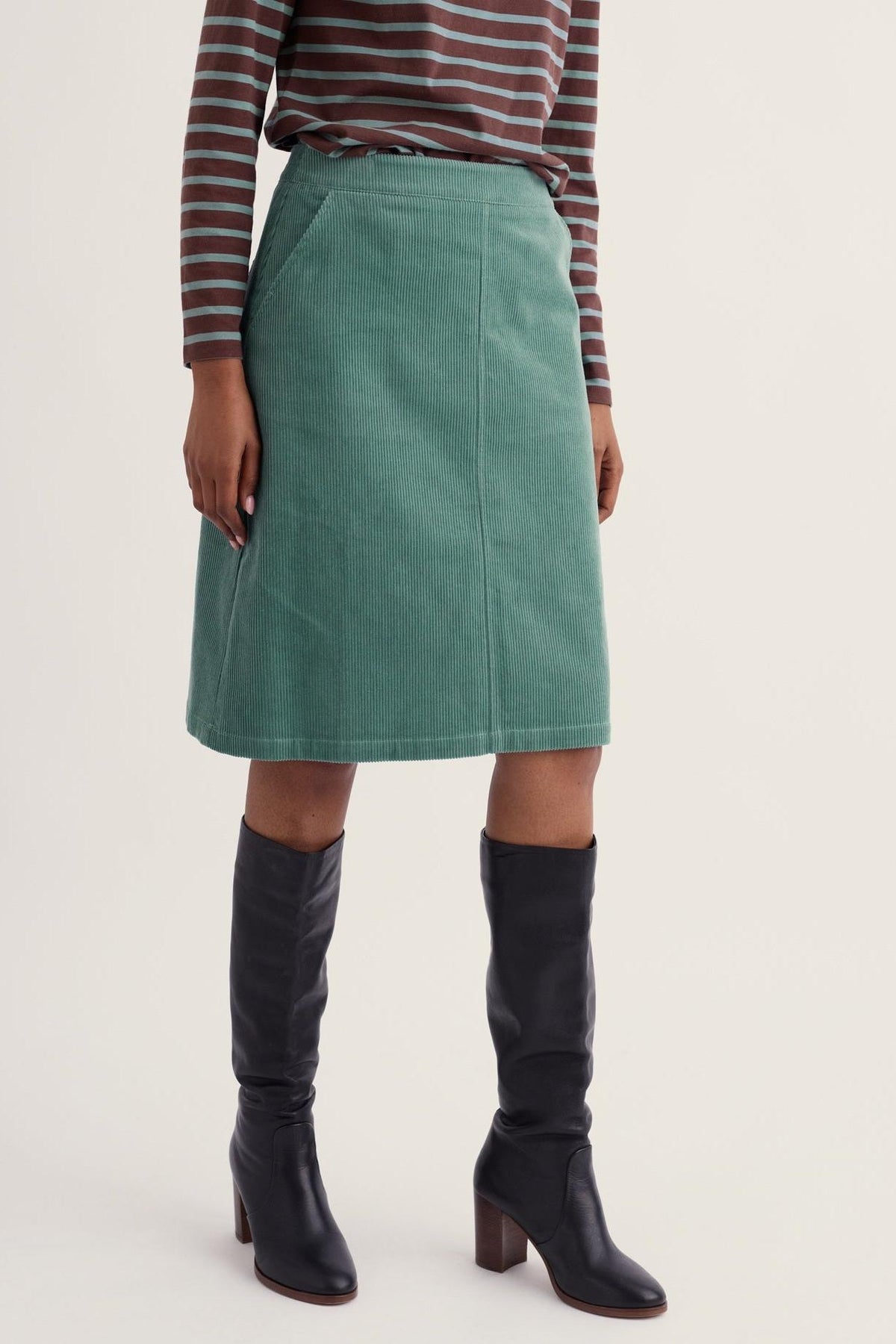 A woman wearing a knee-length teal corduroy skirt from Seasalt Cornwall, paired with black leather boots.