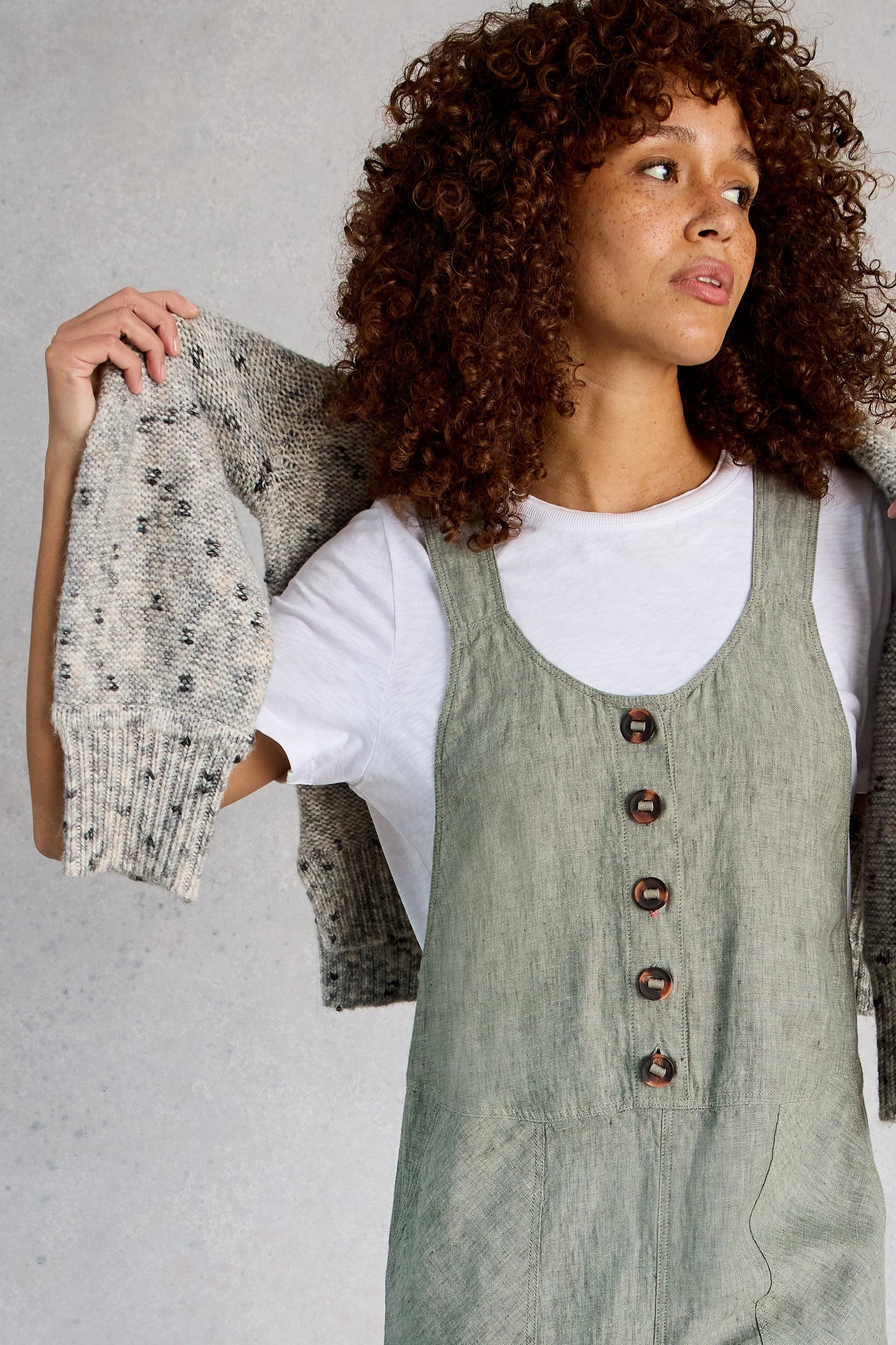 Woman wearing a green White Stuff dungarees over a white shirt, holding a patterned sweater against a neutral background