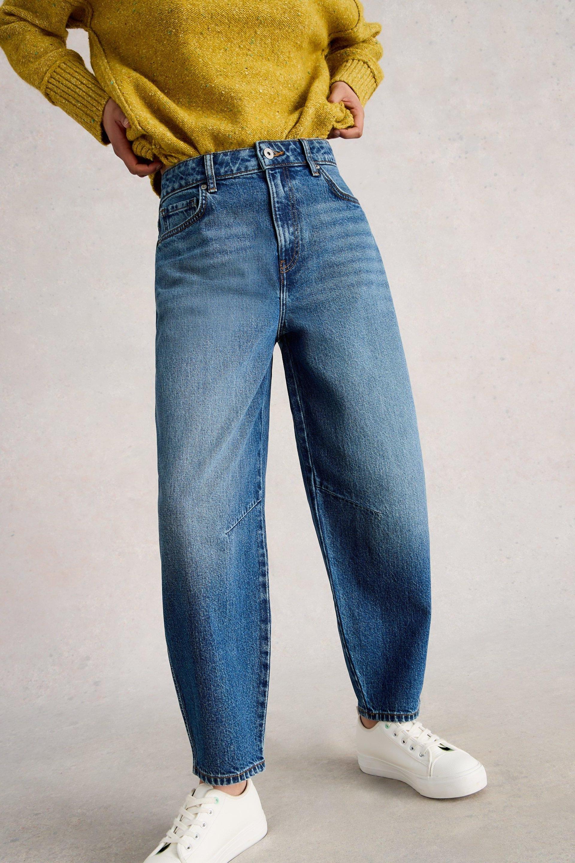 A woman wearing blue mid-denim barrel leg jeans and a yellow sweater against a neutral background.
