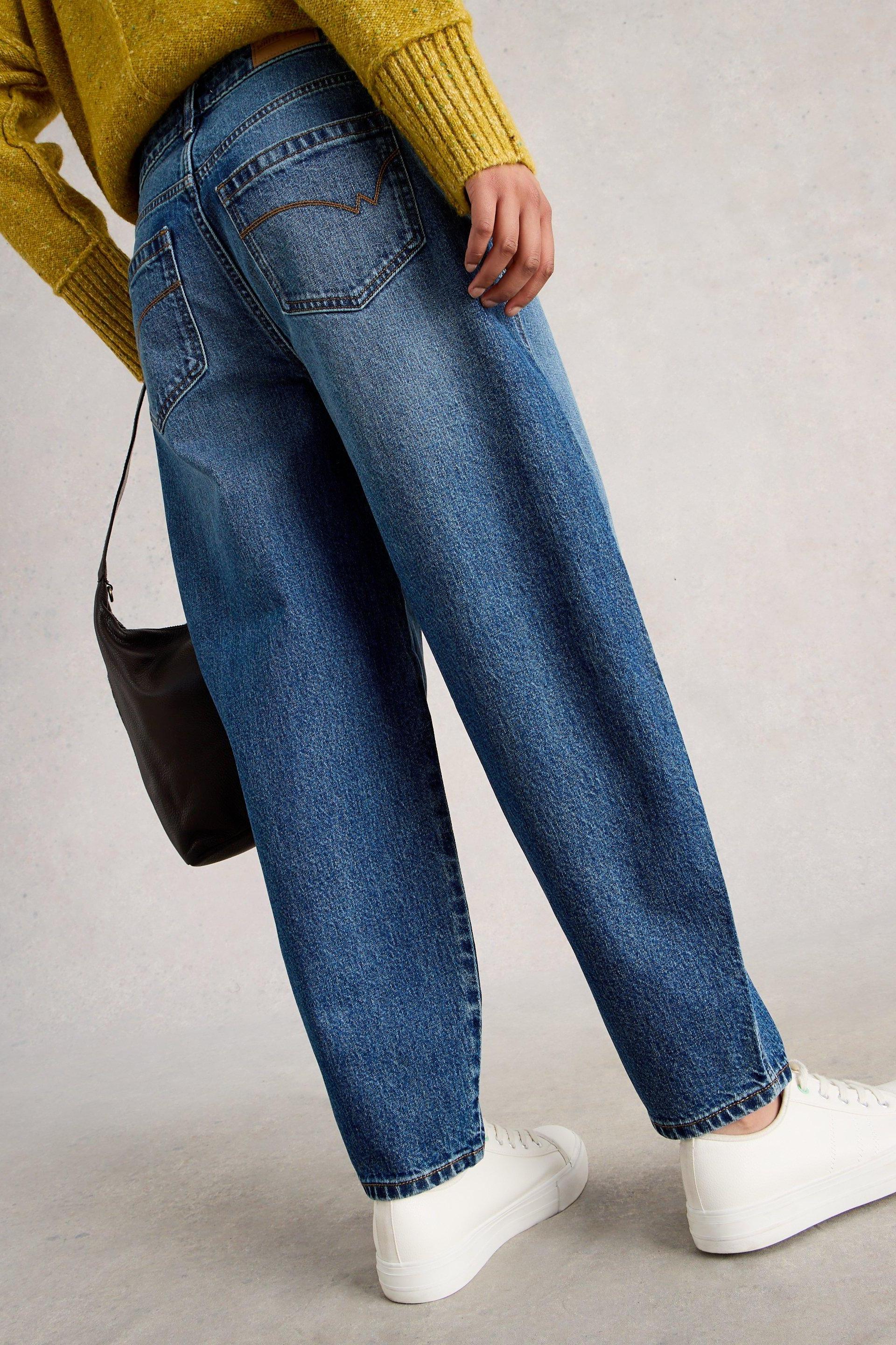 A woman wearing mid-denim barrel leg jeans with a mustard yellow sweater and white sneakers.