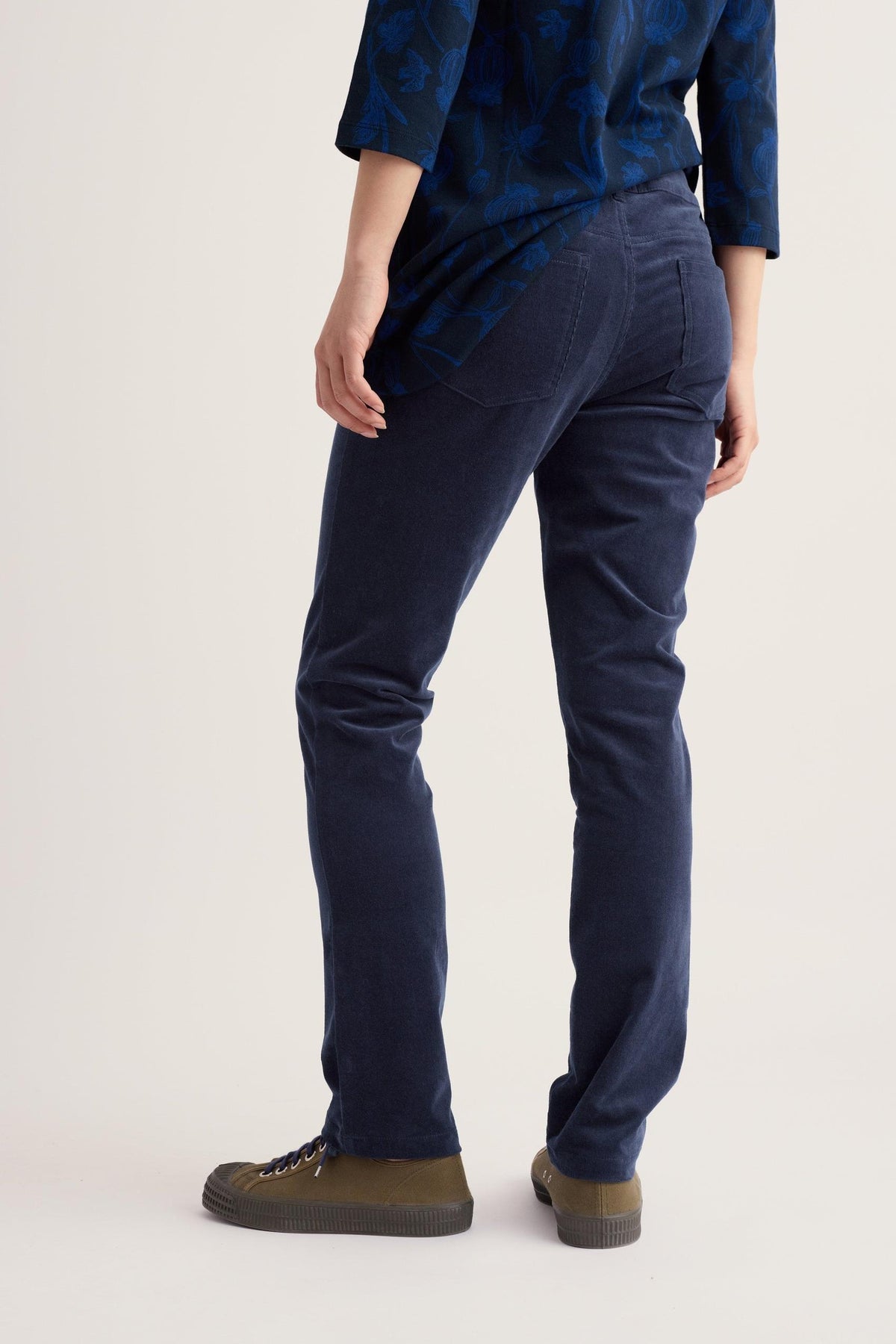 Rear view of a woman wearing navy corduroy trousers and a patterned blue top against a plain background.